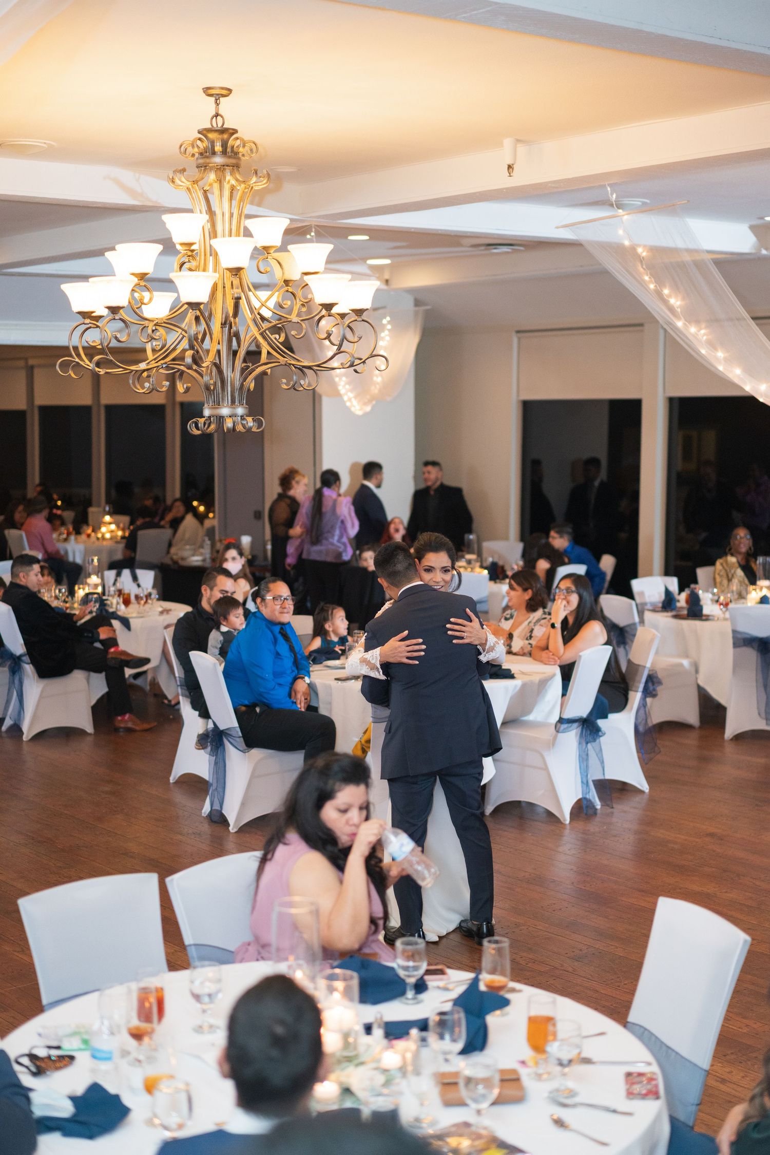 Elegant reception hall with crystal chandeliers and white-covered chairs set for wedding dinner.