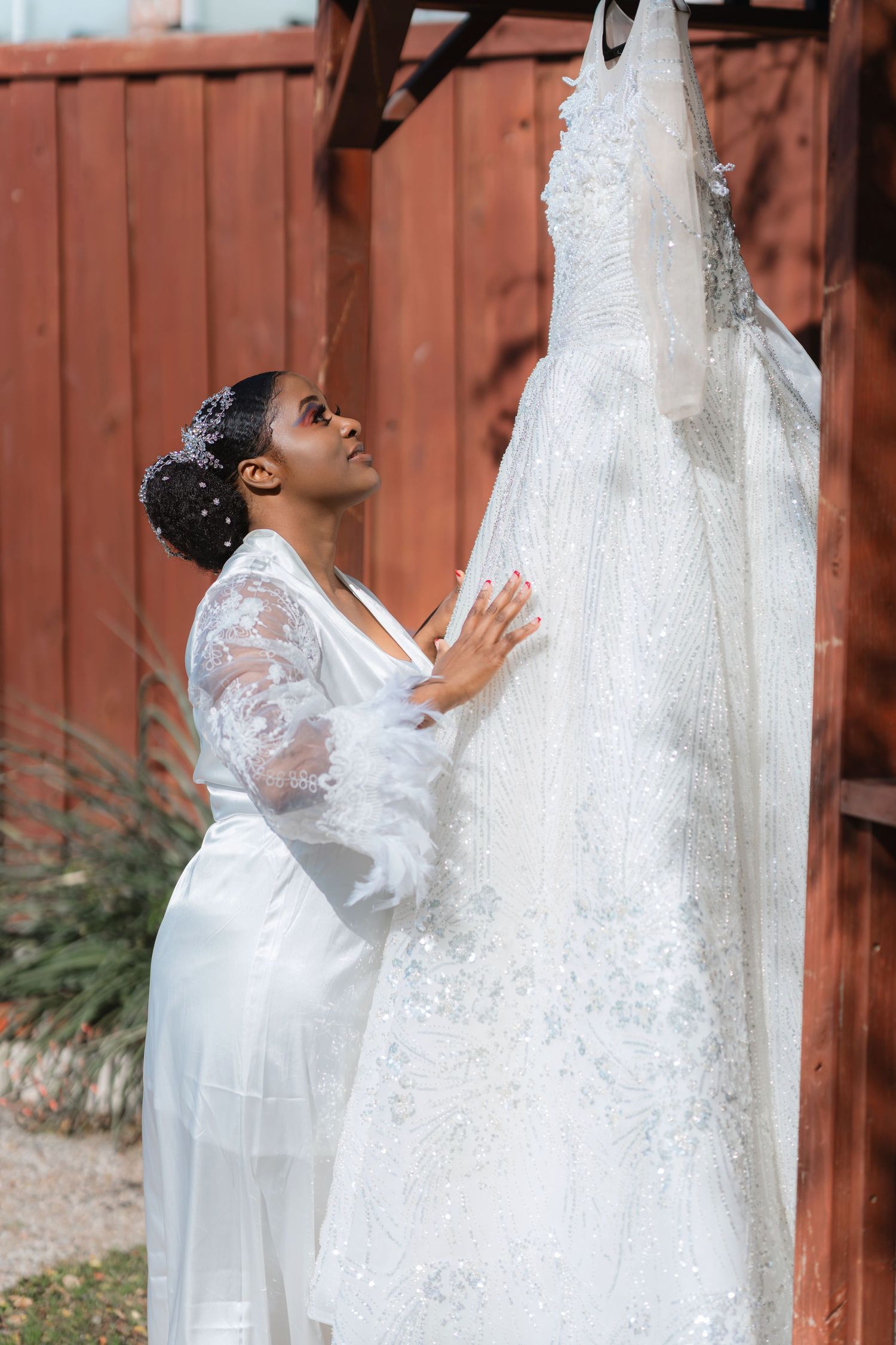 Bride in white robe admiring wedding dress hanging outdoors by red barn.