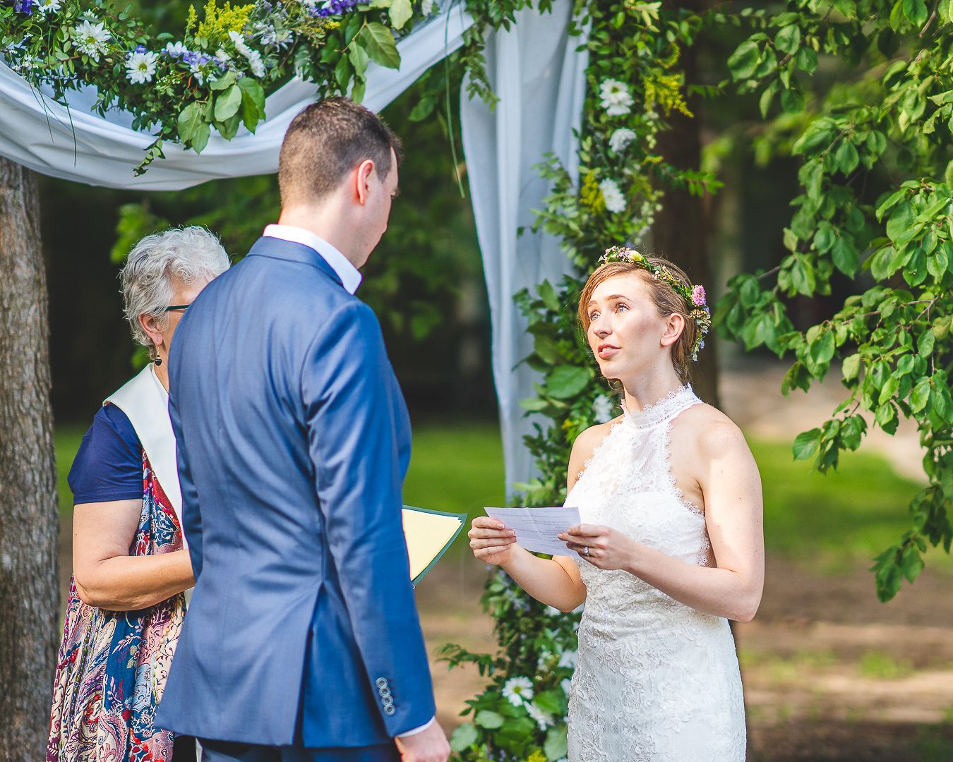 An outdoor wedding ceremony taking place under trees with white fabric decorations.