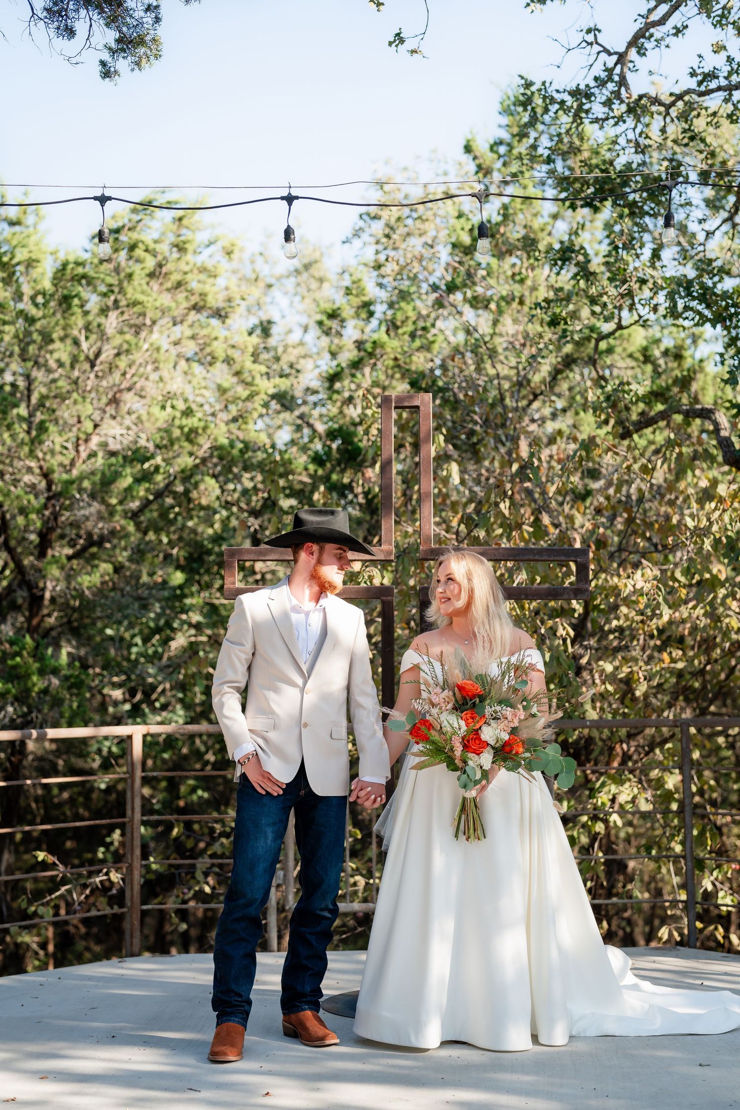 A couple in wedding attire shares an intimate moment on an outdoor wooden deck surrounded by trees.