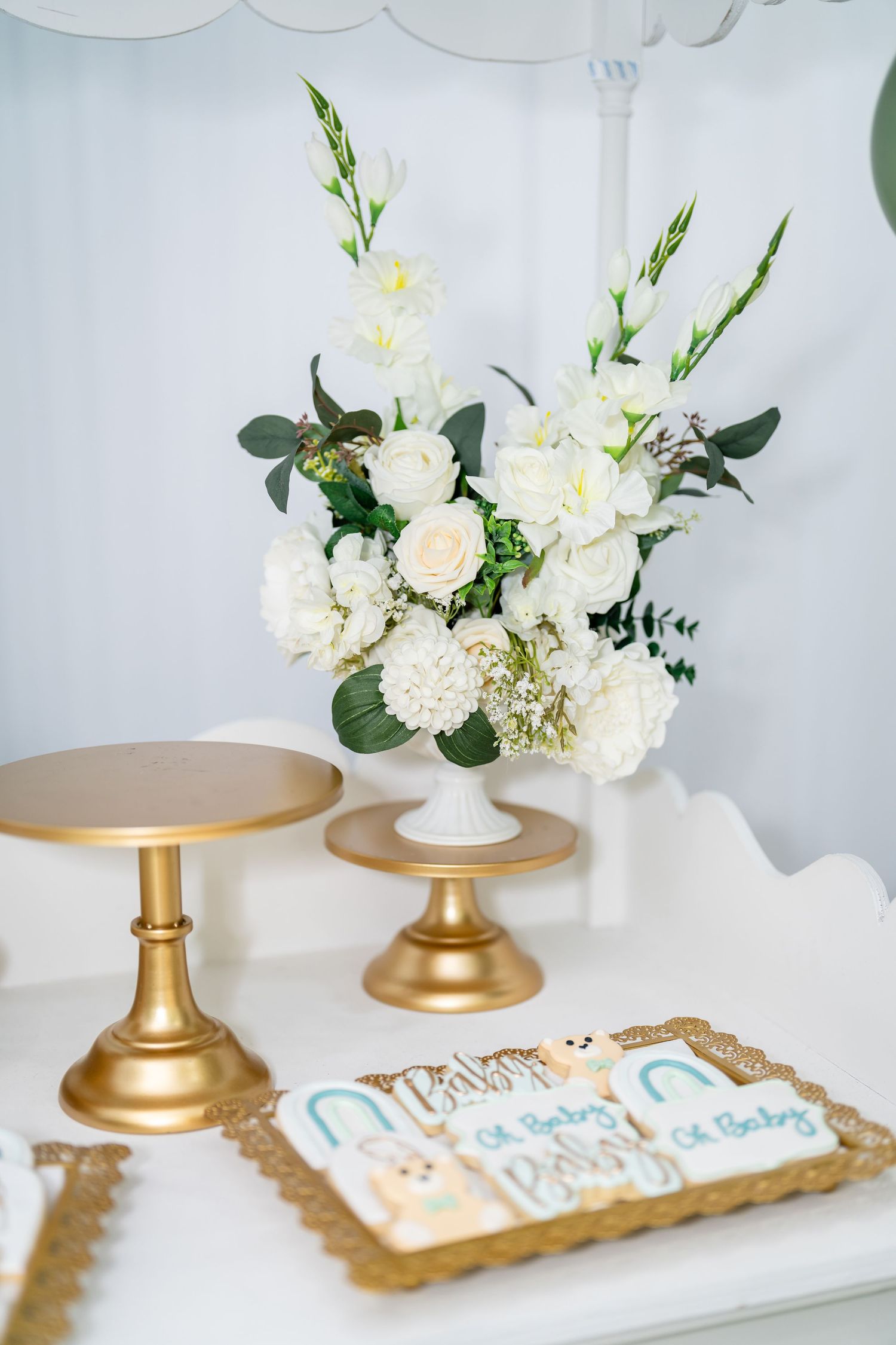 Elegant gold cake stands with white floral arrangements and decorated cookies displayed on a white surface.