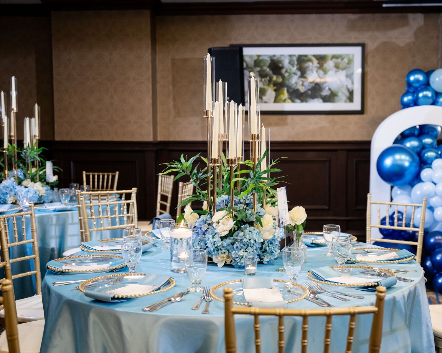 Elegant wedding reception tables decorated with blue linens, white flowers, and gold accents in a banquet hall setting.