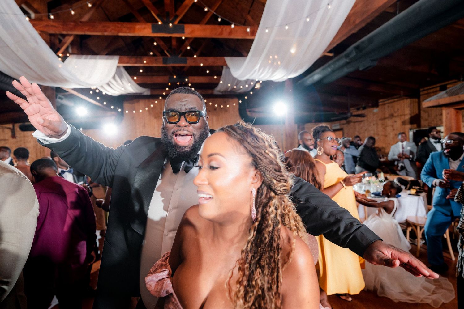 Wedding guests dance and celebrate joyfully on a reception dance floor with festive lighting overhead.