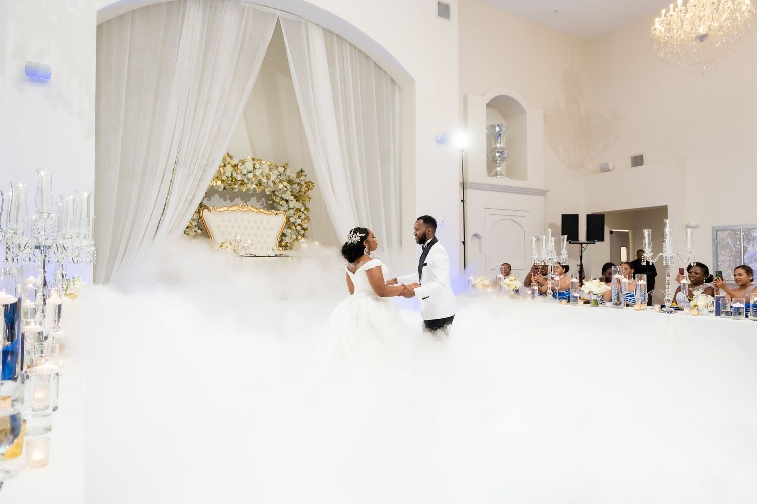 Spacious white wedding venue with draped fabric, chandeliers and guest tables arranged for celebration.