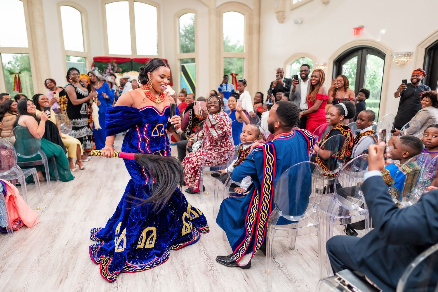 Wedding guests dancing and celebrating in a bright reception hall with arched windows.