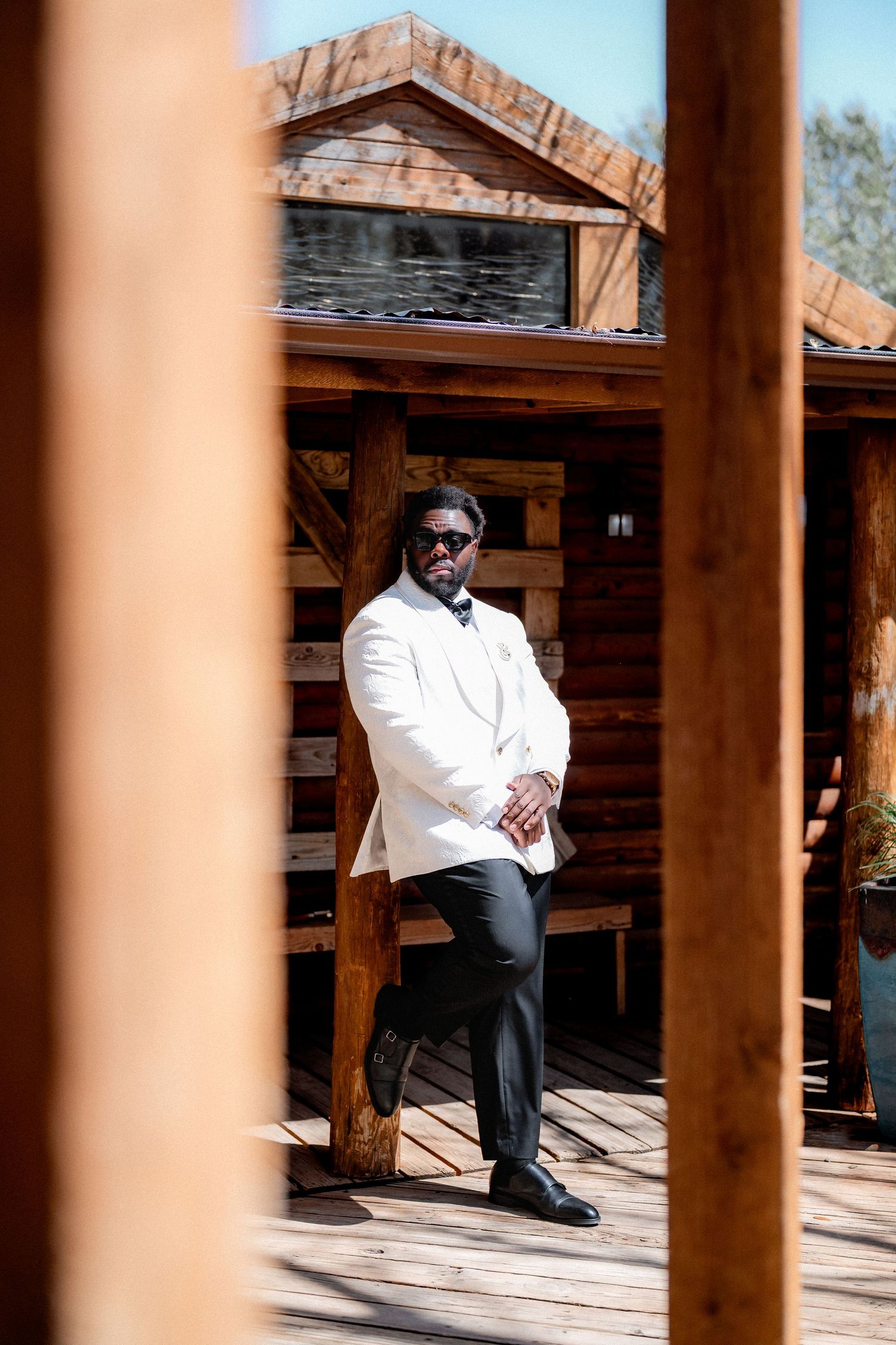Groom in white suit jacket and black pants sits thoughtfully on rustic wooden porch with wooden pillars.