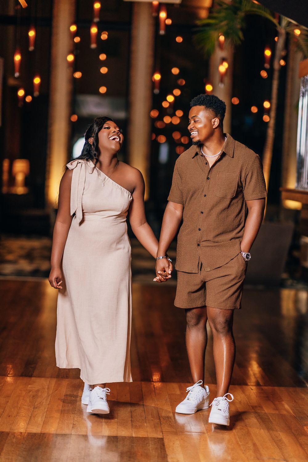 Couple walking hand in hand in brown and beige outfits on a wooden floor with twinkling lights.