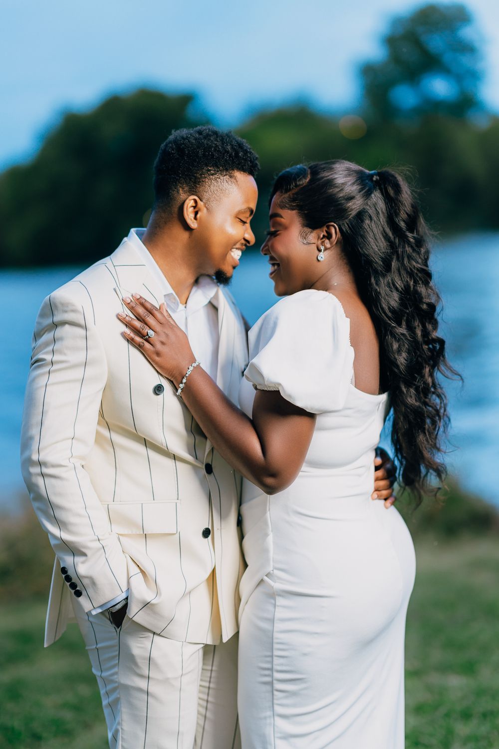 Romantic couple in white outfits share intimate moments by a waterfront during a sunset photoshoot.