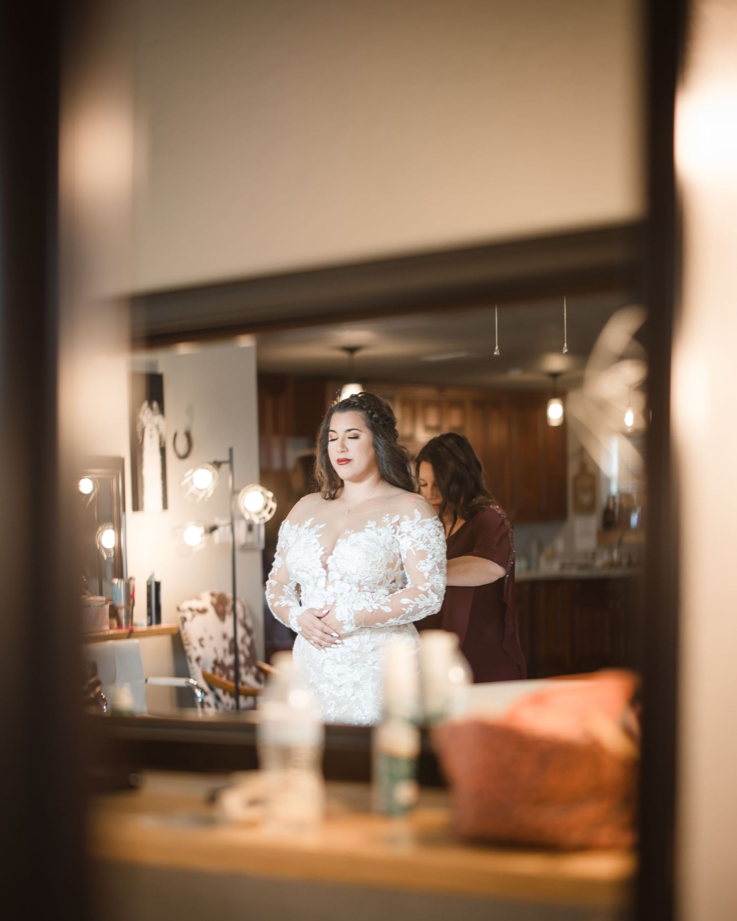 A bride in a white lace wedding dress is reflected in a mirror during wedding preparations.