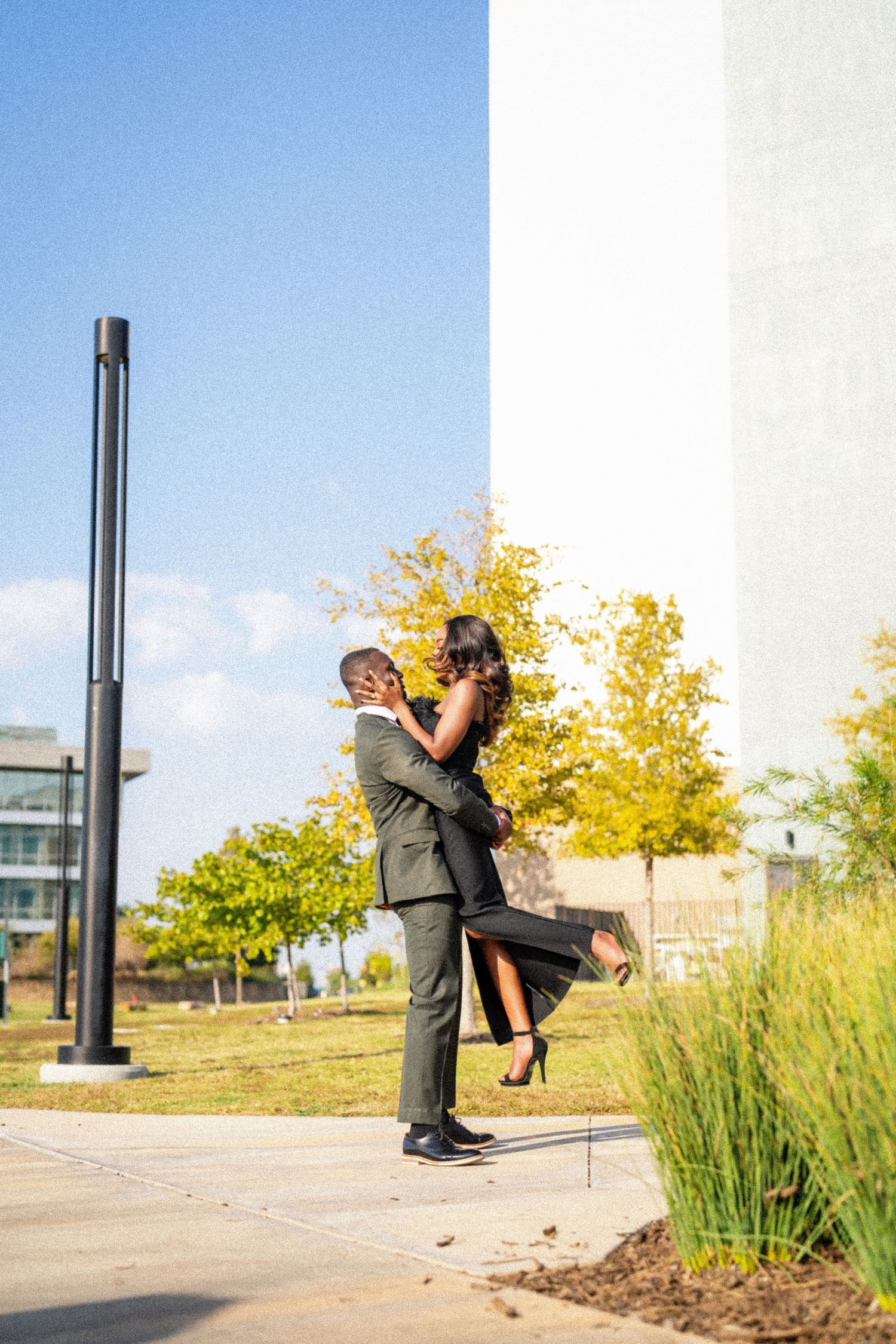 A couple in formal wear share a romantic lift pose against a blue sky and autumn trees.