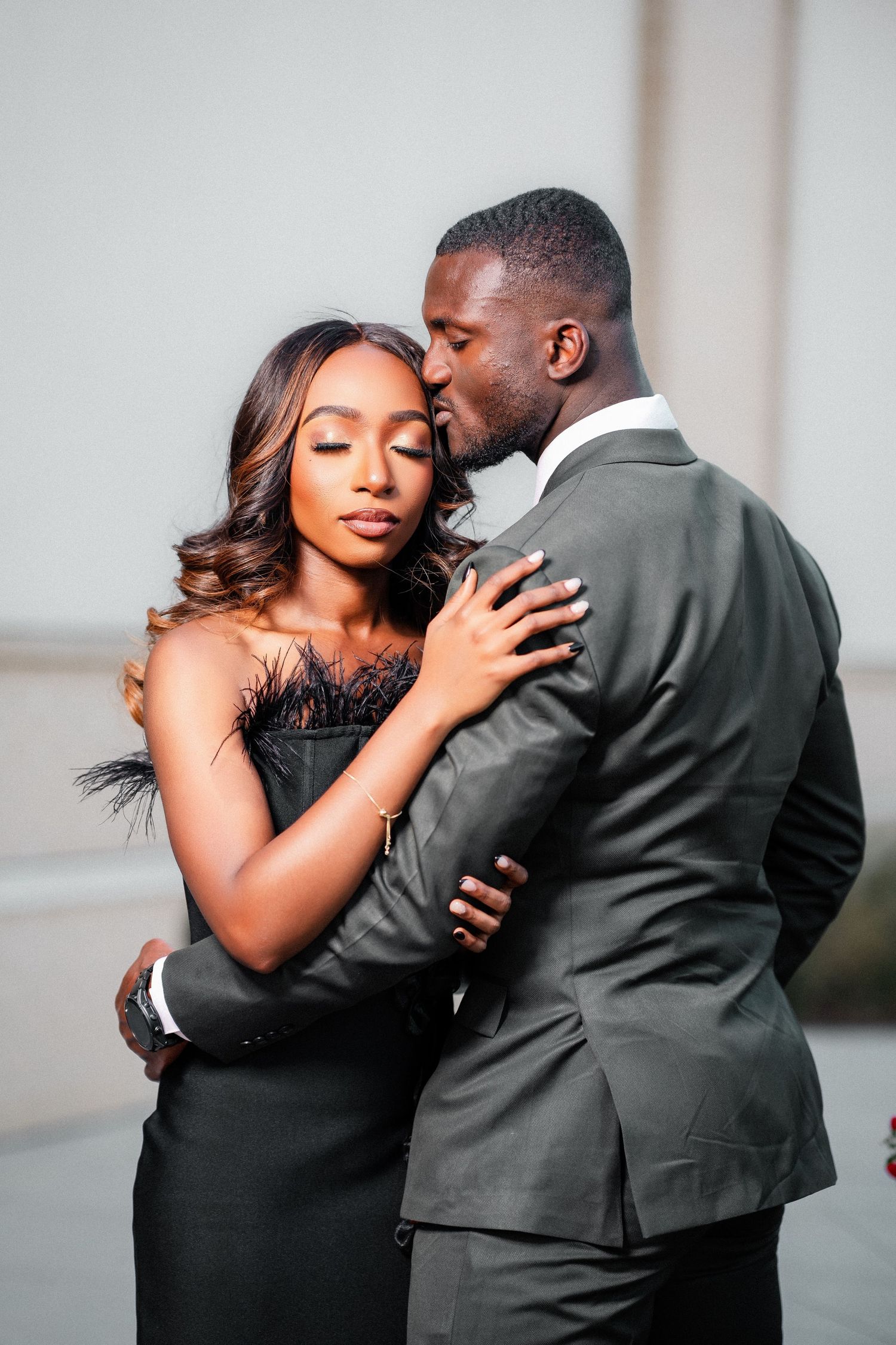 A couple in black and grey formal wear embrace tenderly against a neutral background.