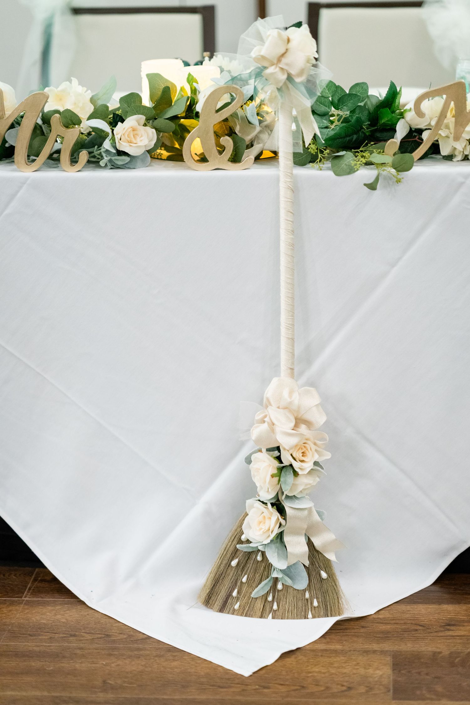 Decorative wedding broom with white roses and eucalyptus leaves against white tablecloth backdrop.