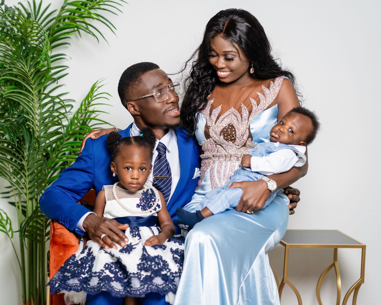 A family in coordinating blue and white outfits poses together against a white backdrop with tropical greenery.
