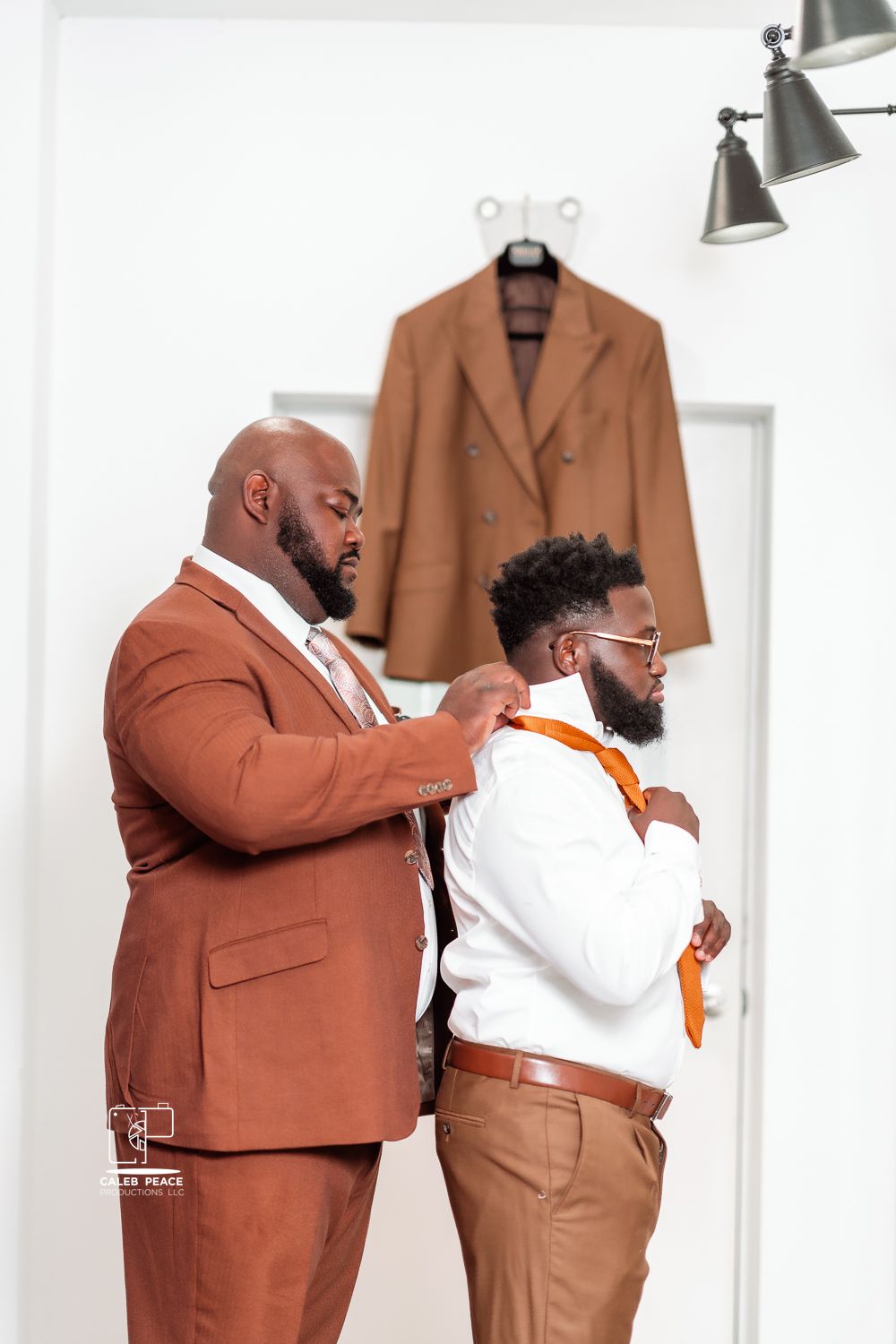 Two men in brown suits helping each other get dressed and adjust their formal attire.