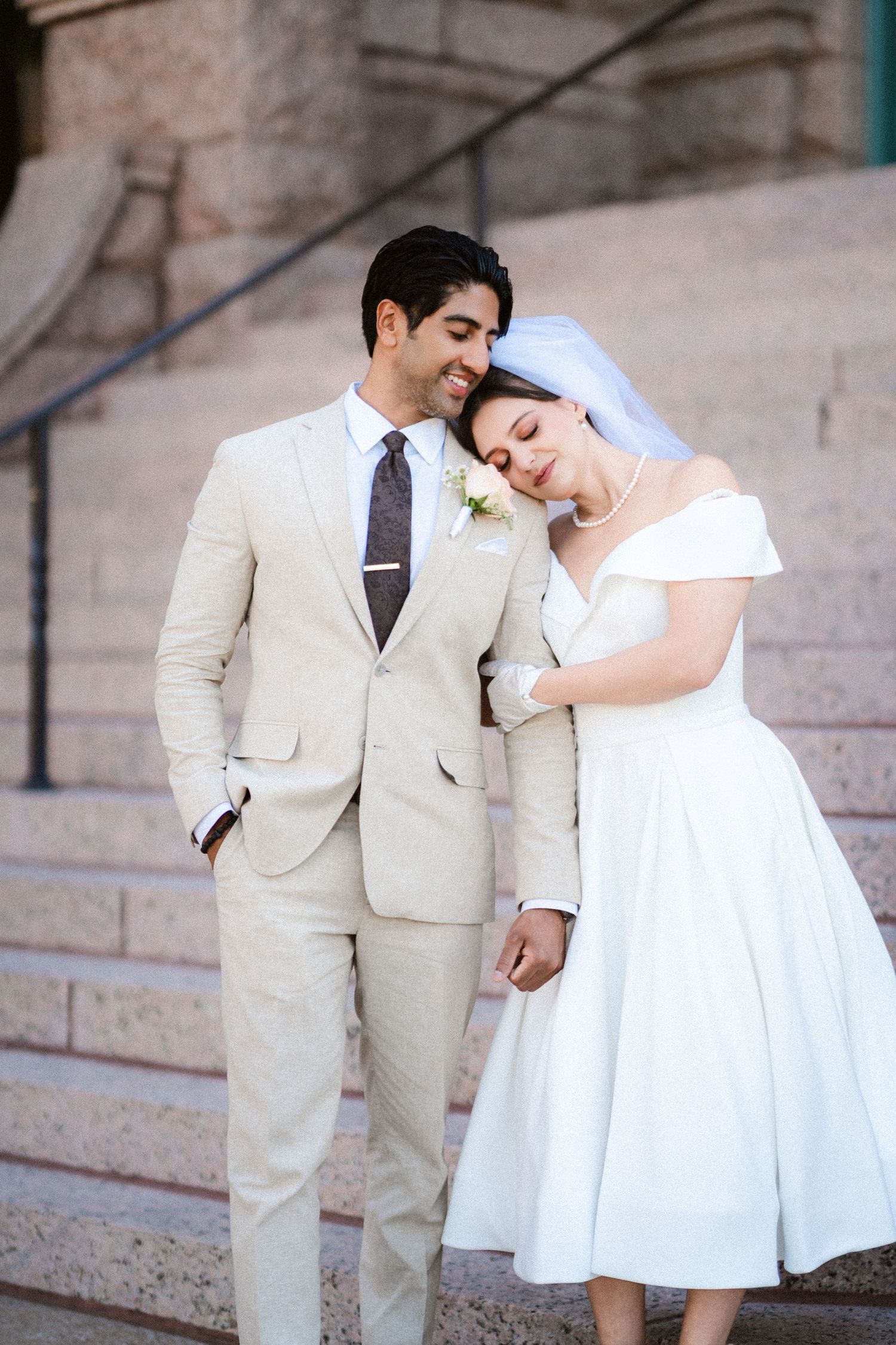 A romantic couple in white and beige formal attire pose playfully on stone steps during a wedding photoshoot.