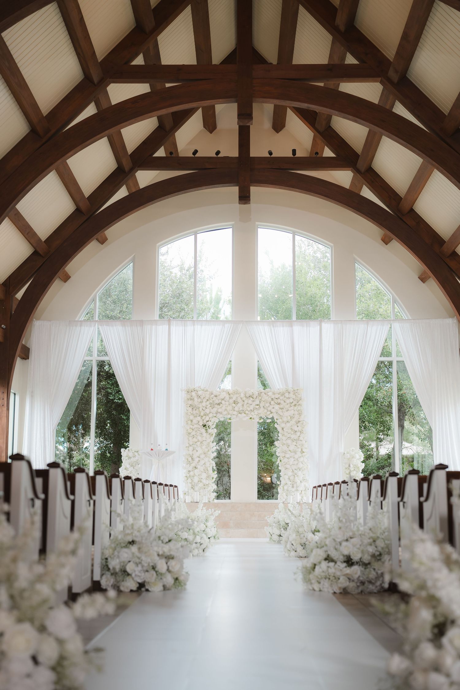 Elegant wedding venue interior with white floral arrangements and sheer curtains along arched wooden beam ceiling.