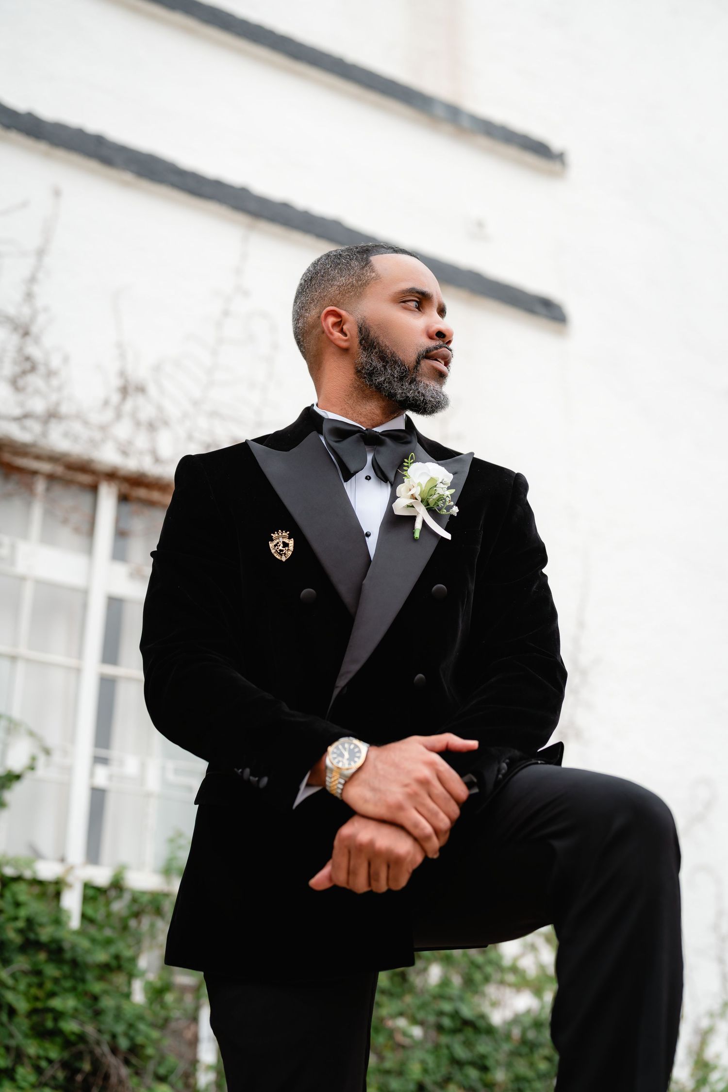 Groom in classic black tuxedo and bowtie poses thoughtfully against white building exterior with boutonniere.