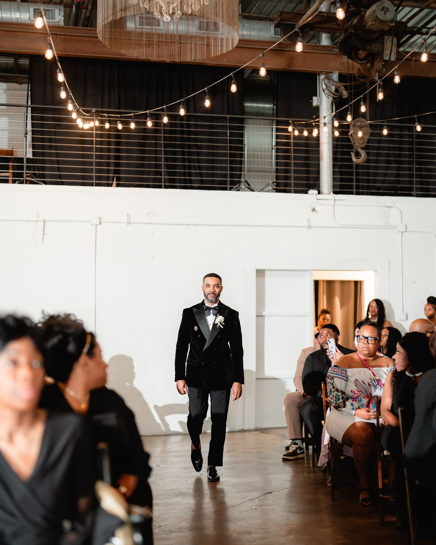 Groom walking down aisle in industrial venue with string lights and modern decor.