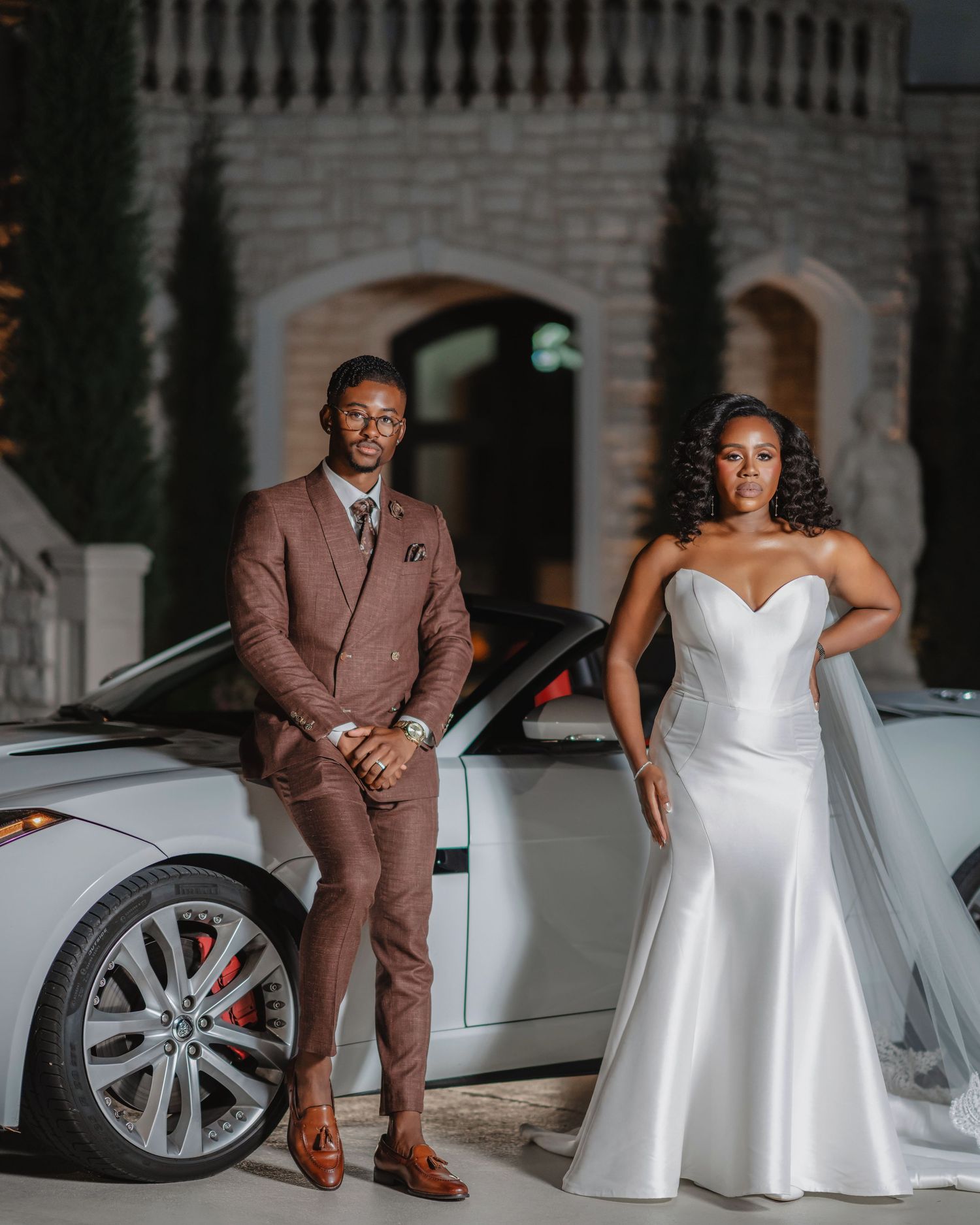 A couple in wedding attire poses together by a white luxury car for elegant wedding photos.