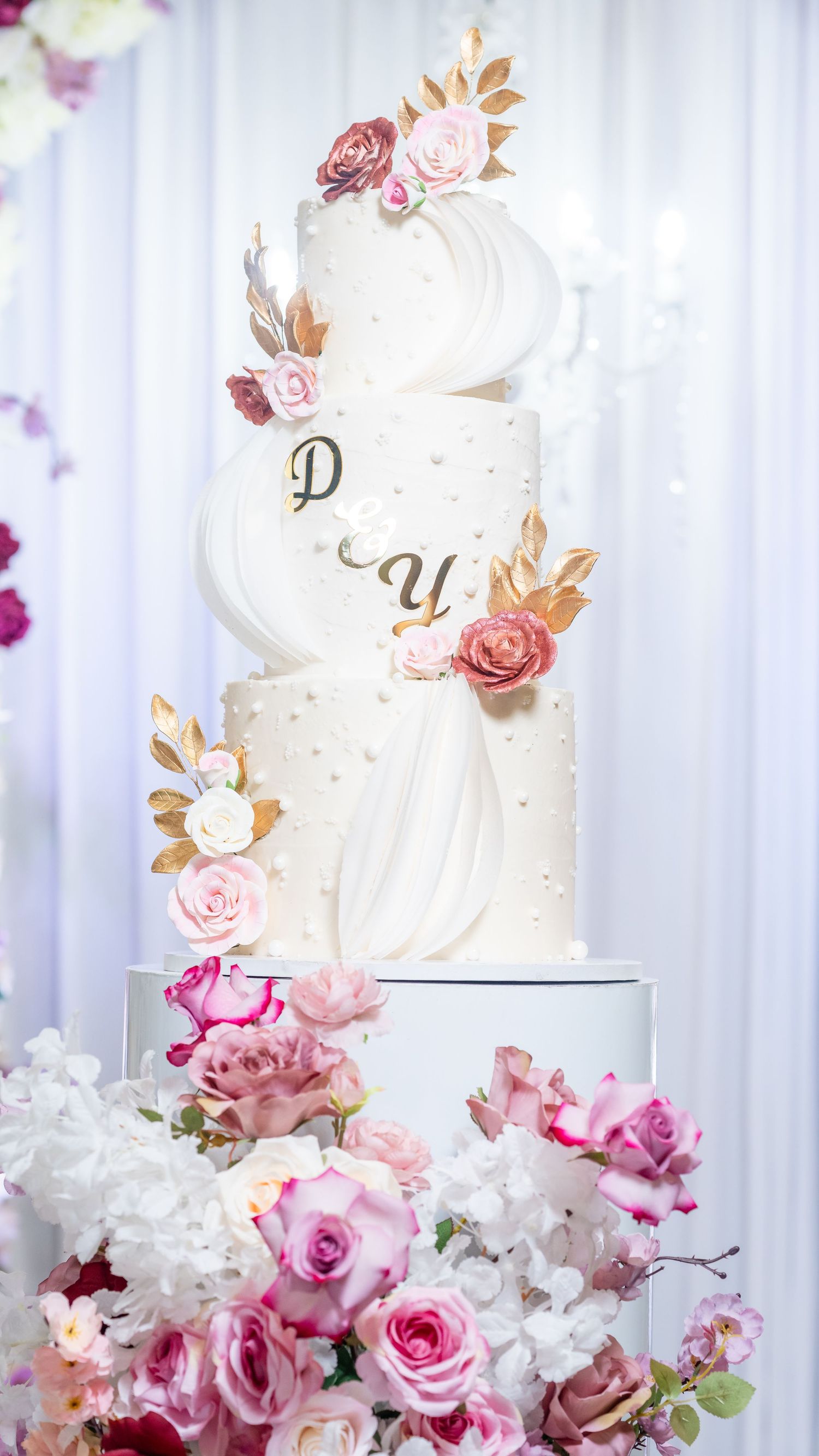 Elegant white wedding cake decorated with pink roses and metallic accents against a light backdrop.