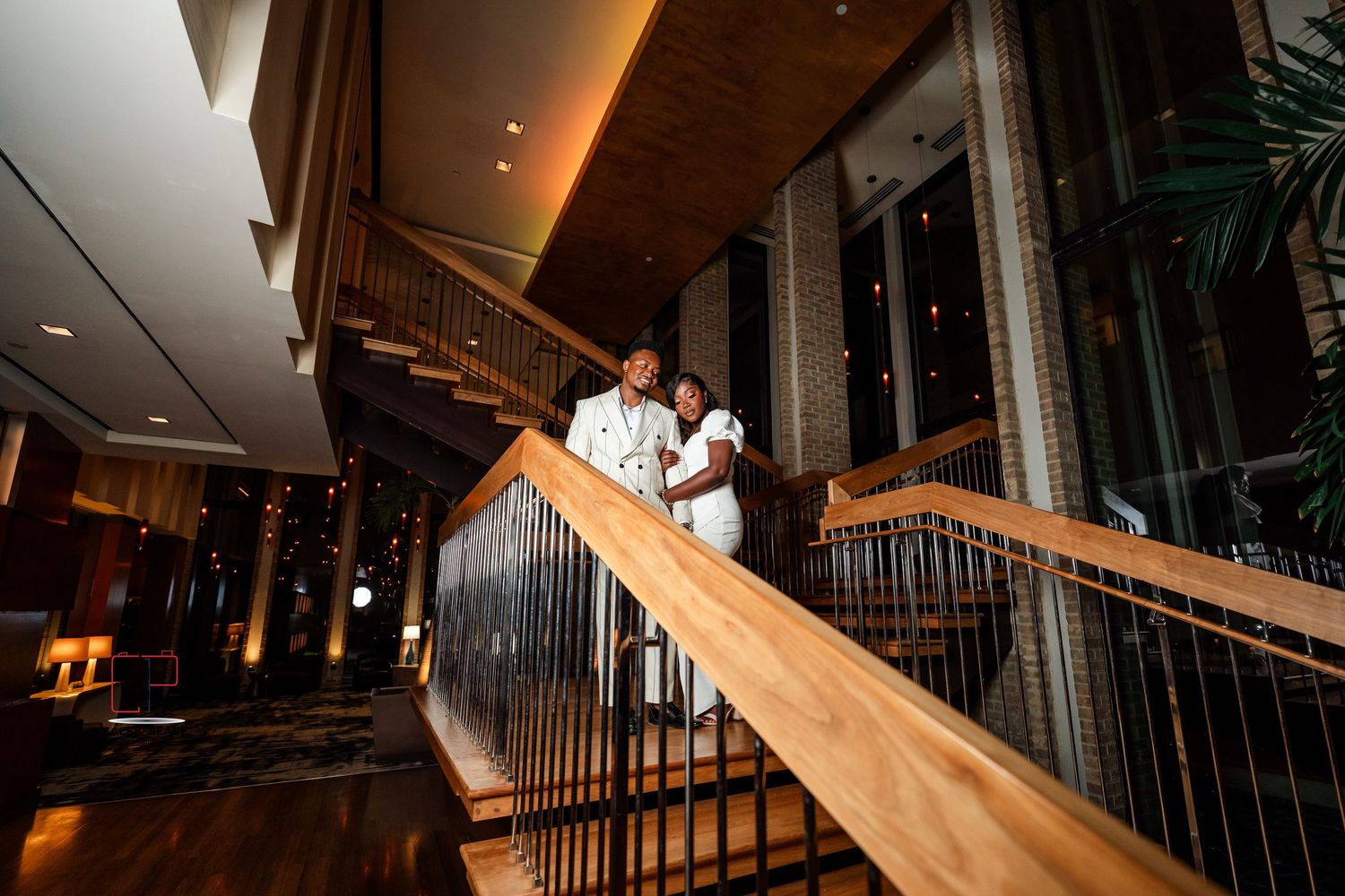 A couple in formal attire stands on an elegant curved wooden staircase with metal railings in a luxurious hotel lobby.