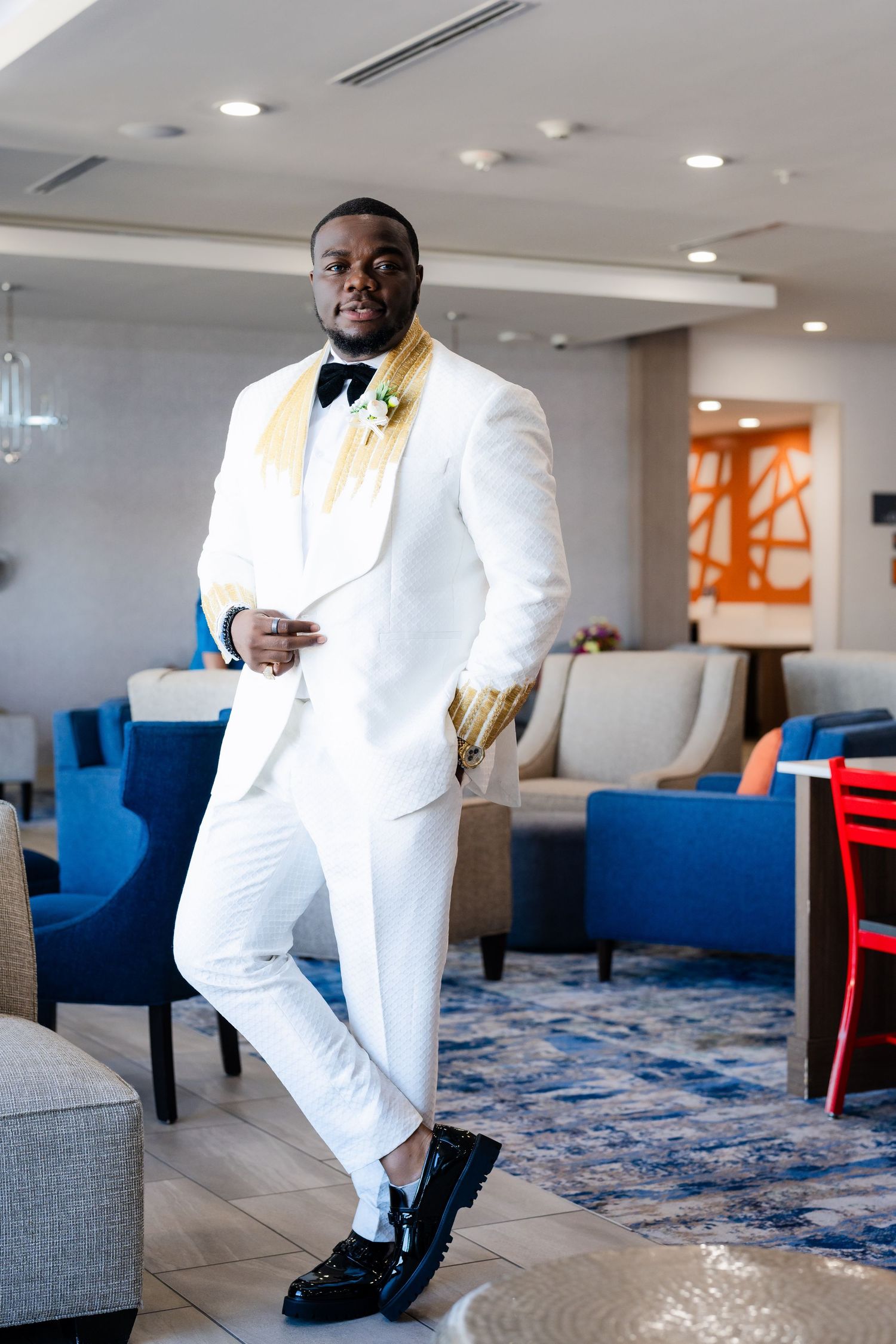 A stylish groom poses in a crisp white tuxedo with gold bow tie in a modern hotel lobby setting.