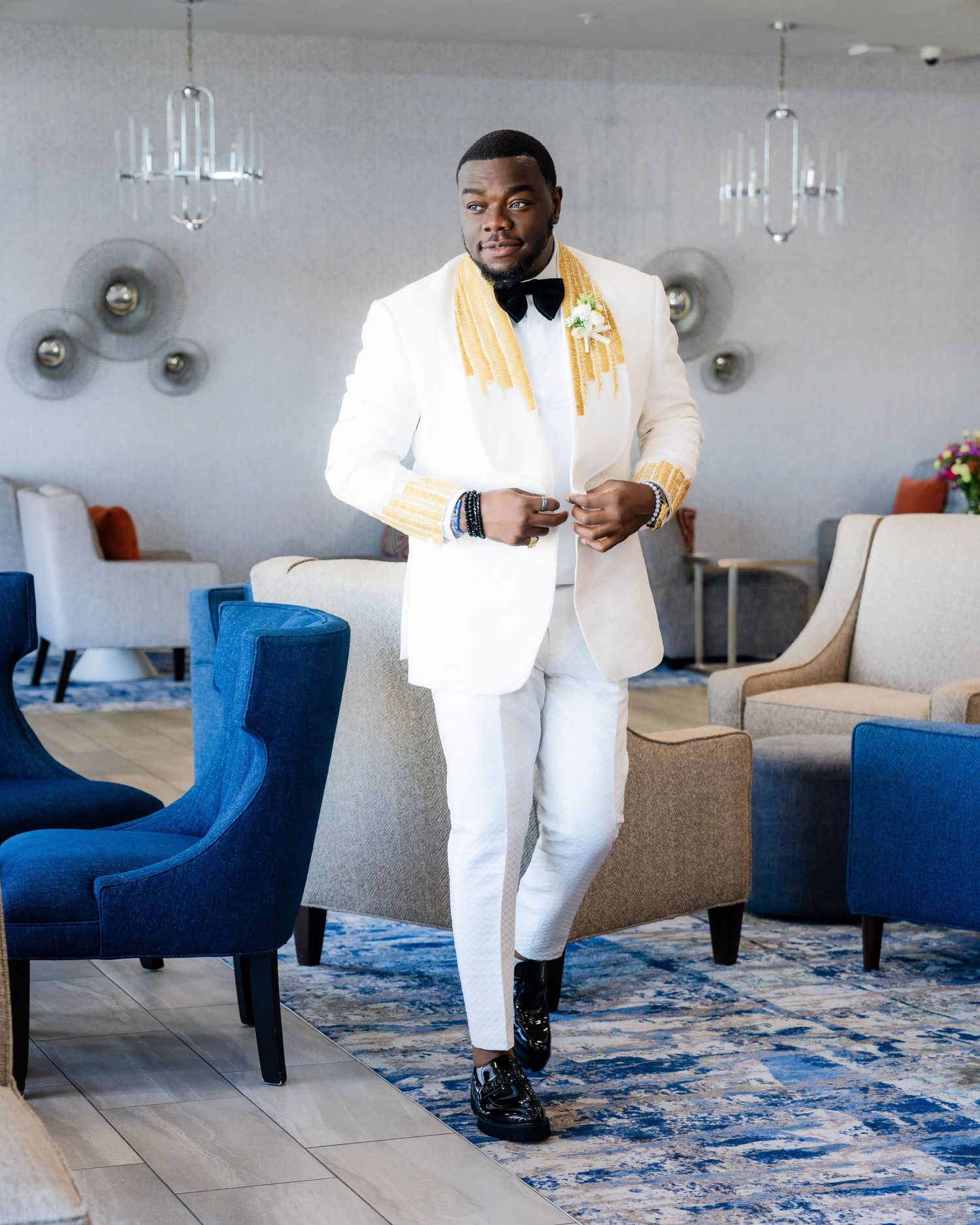 A person in a crisp white tuxedo poses elegantly against a blue velvet chair and patterned rug.