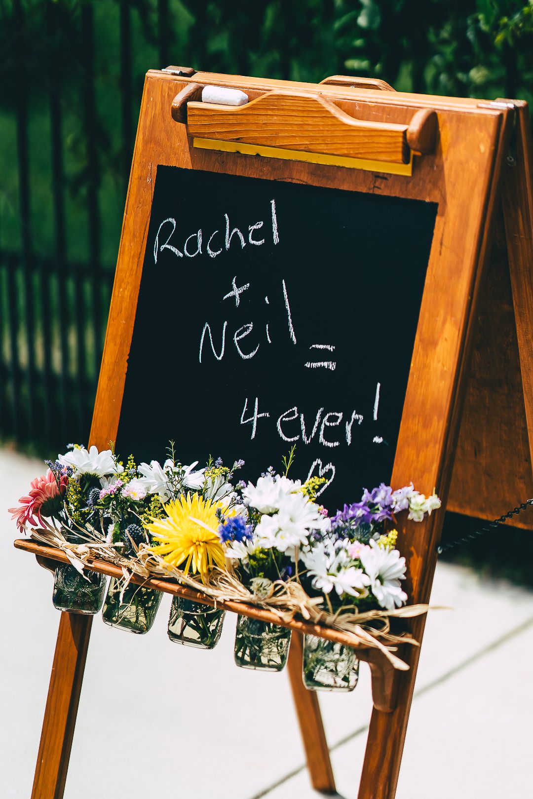 A rustic chalkboard wedding sign decorated with flowers reads 'Rachel + Neil 4ever!' with a heart.