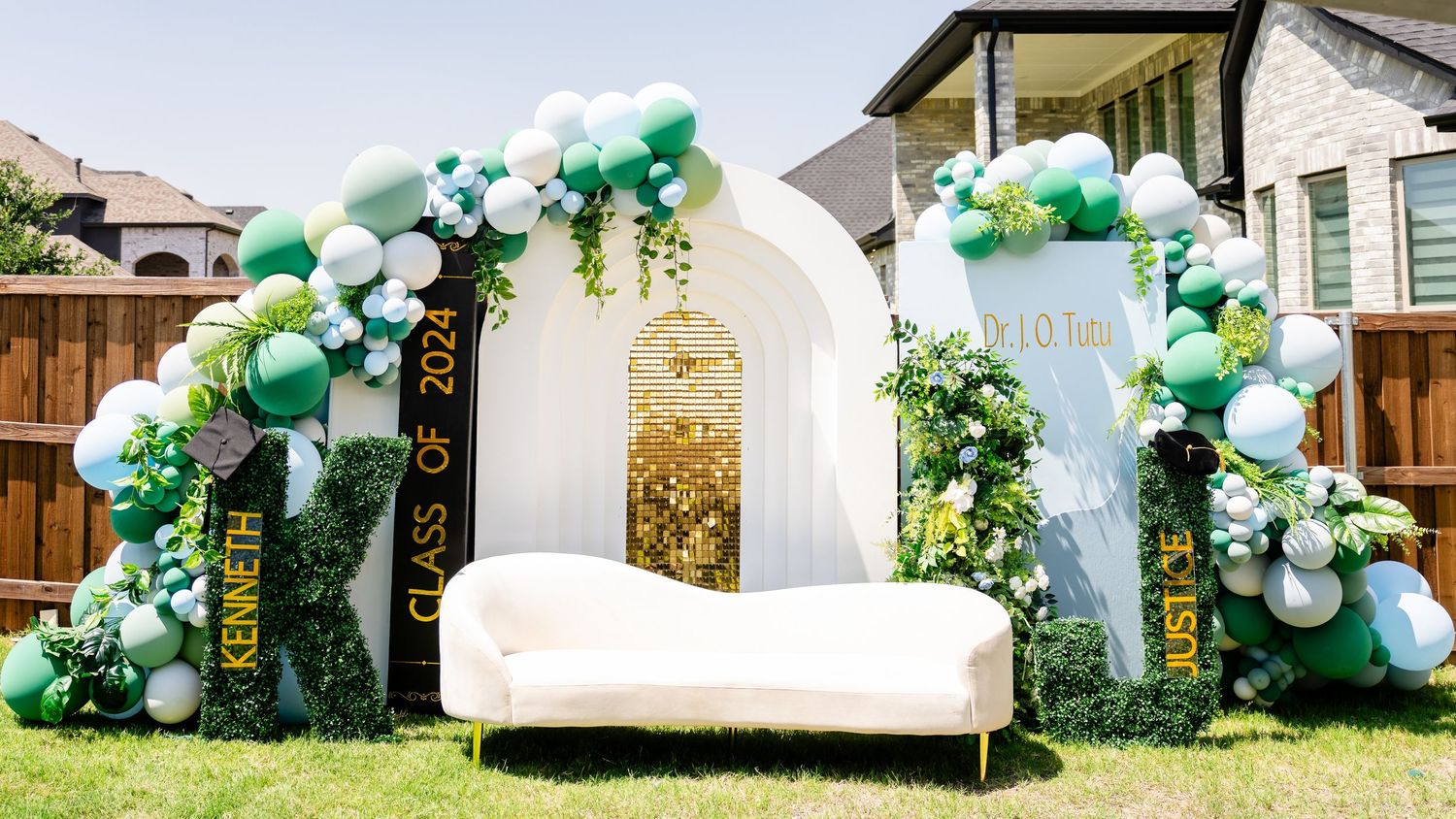 Elegant white sofa between two green and white balloon arches with floral decorations for an outdoor event setup.