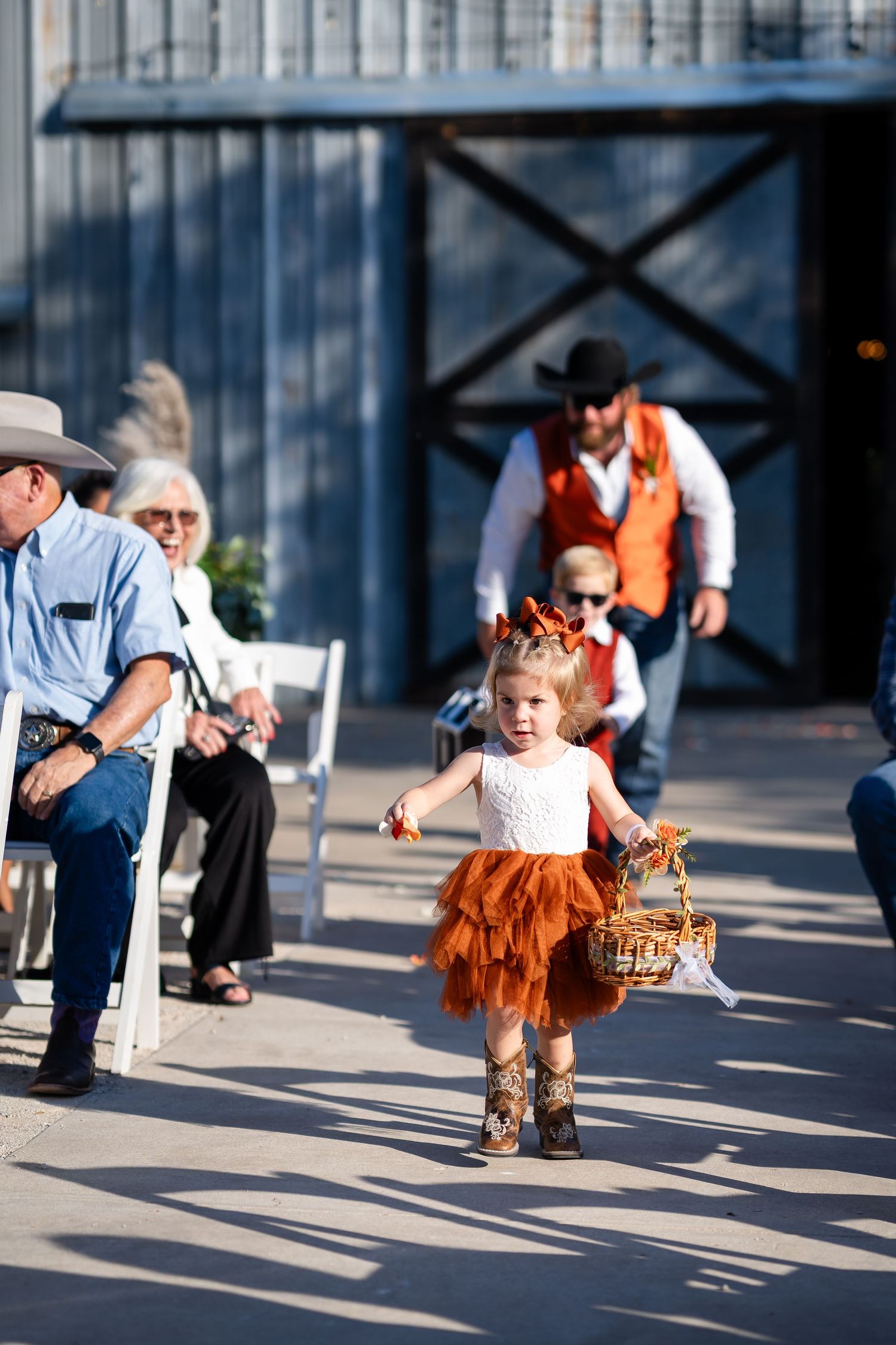 A small child in an orange tutu dress runs playfully across a sunlit patio during a wedding celebration.