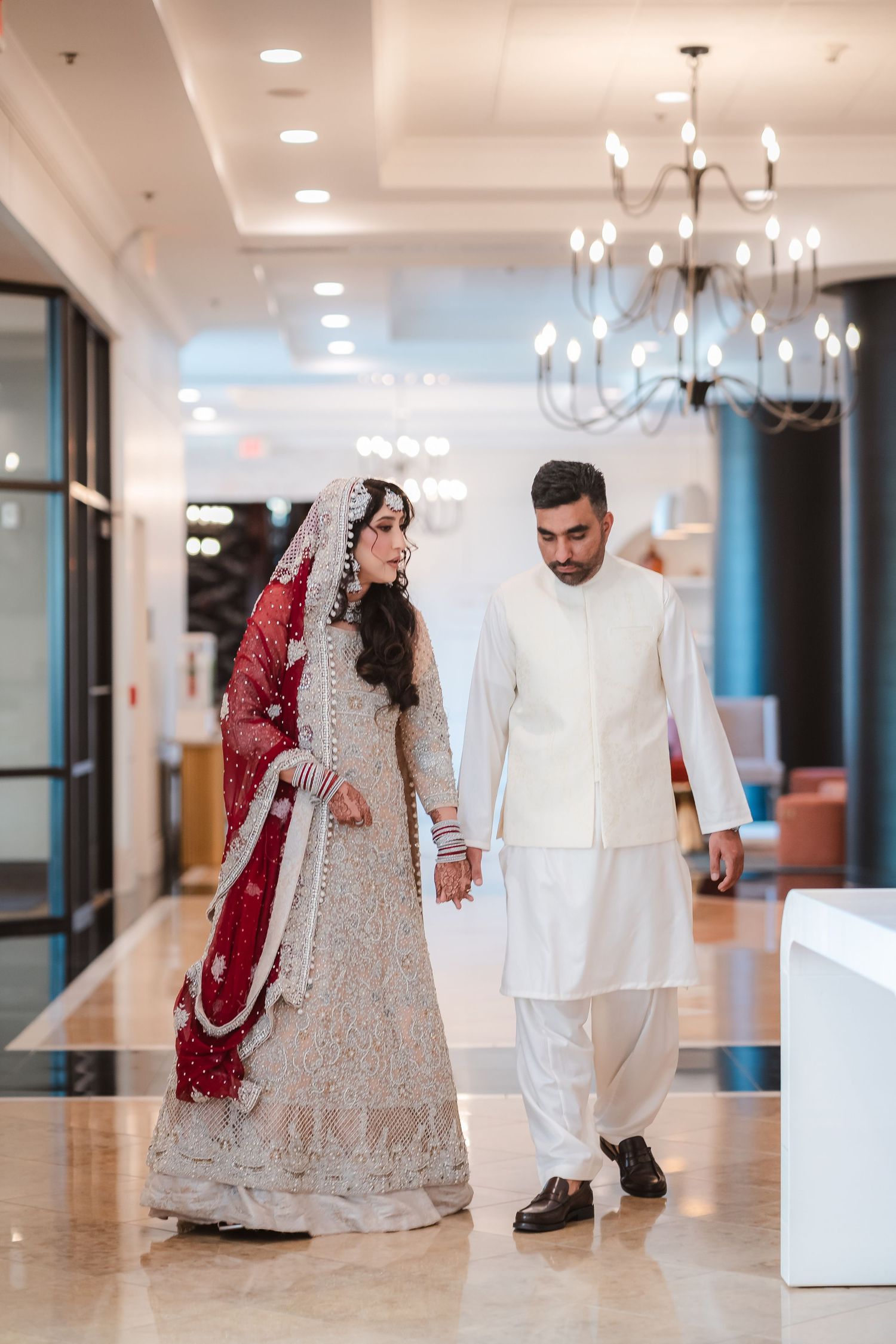 A couple in traditional South Asian wedding attire walks through an elegant hallway with chandeliers.