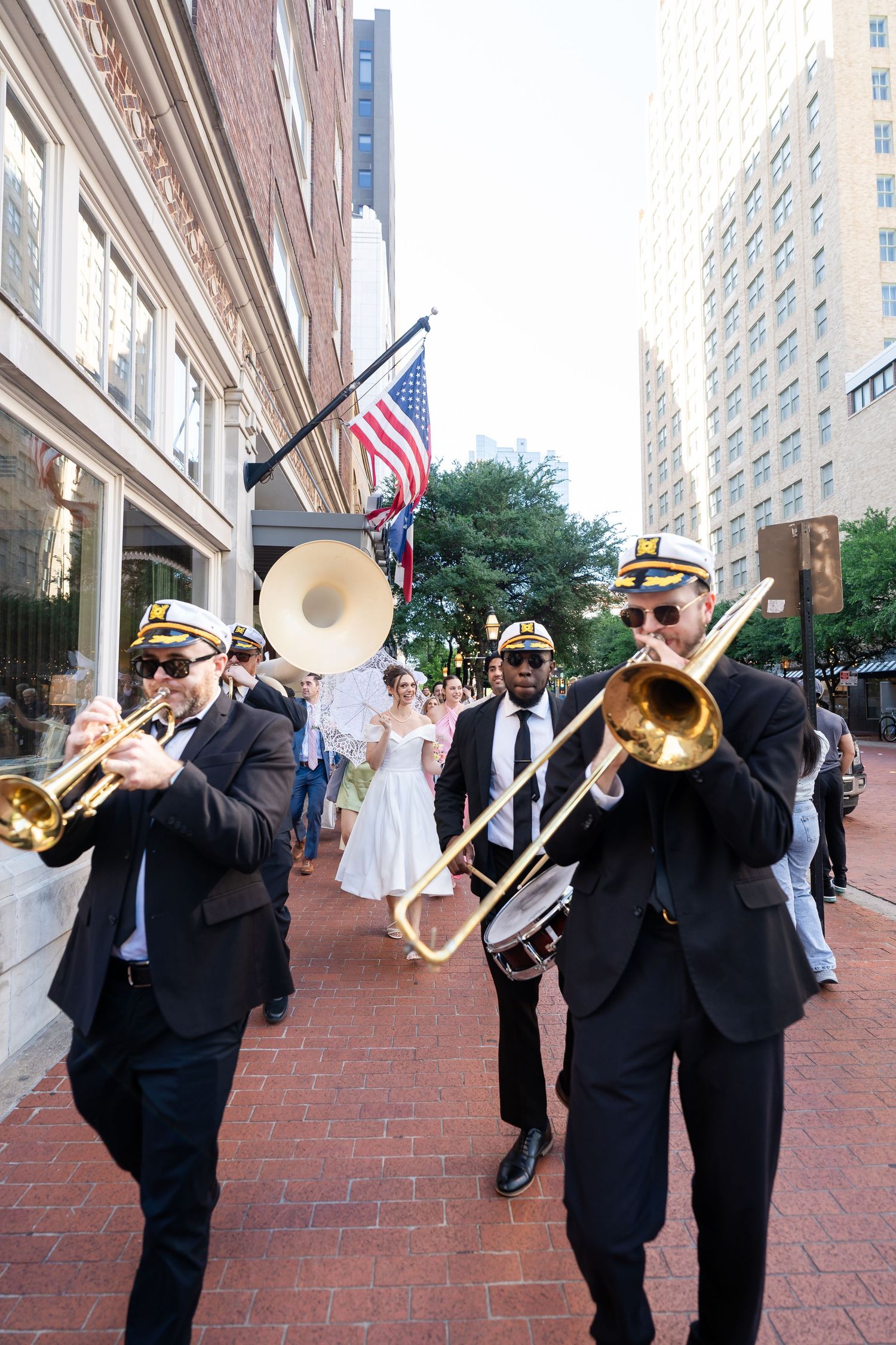 A brass band in formal black attire performs in a line on a brick sidewalk outside a city building.