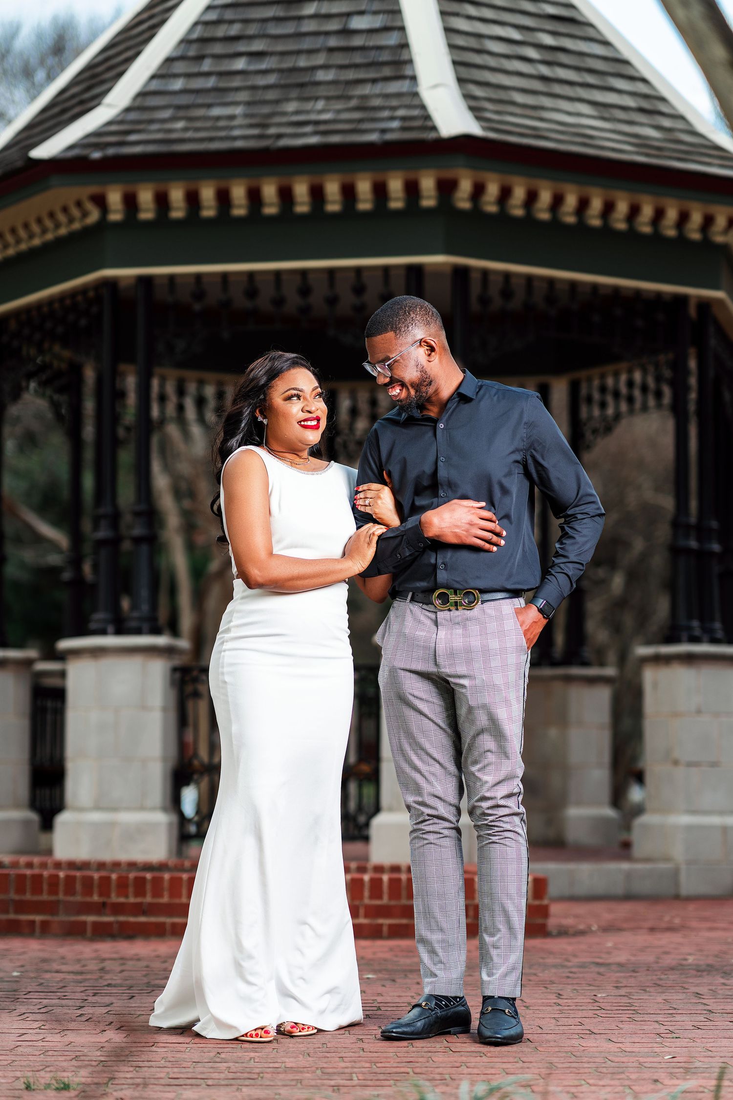 A couple shares laughter while posing together in front of a gazebo on brick pavement.