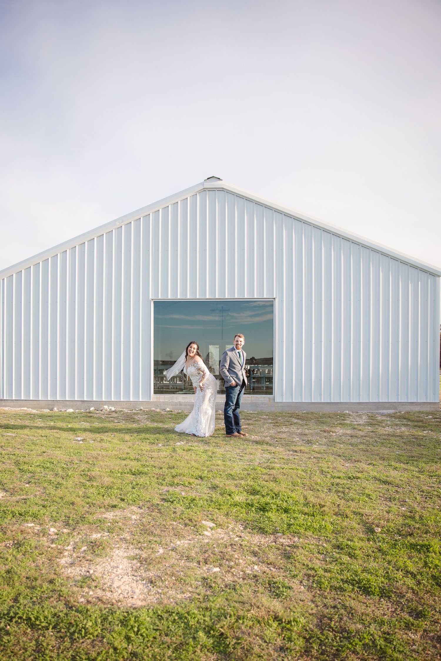 A wedding couple poses in front of a large white metal barn building in a rural setting.