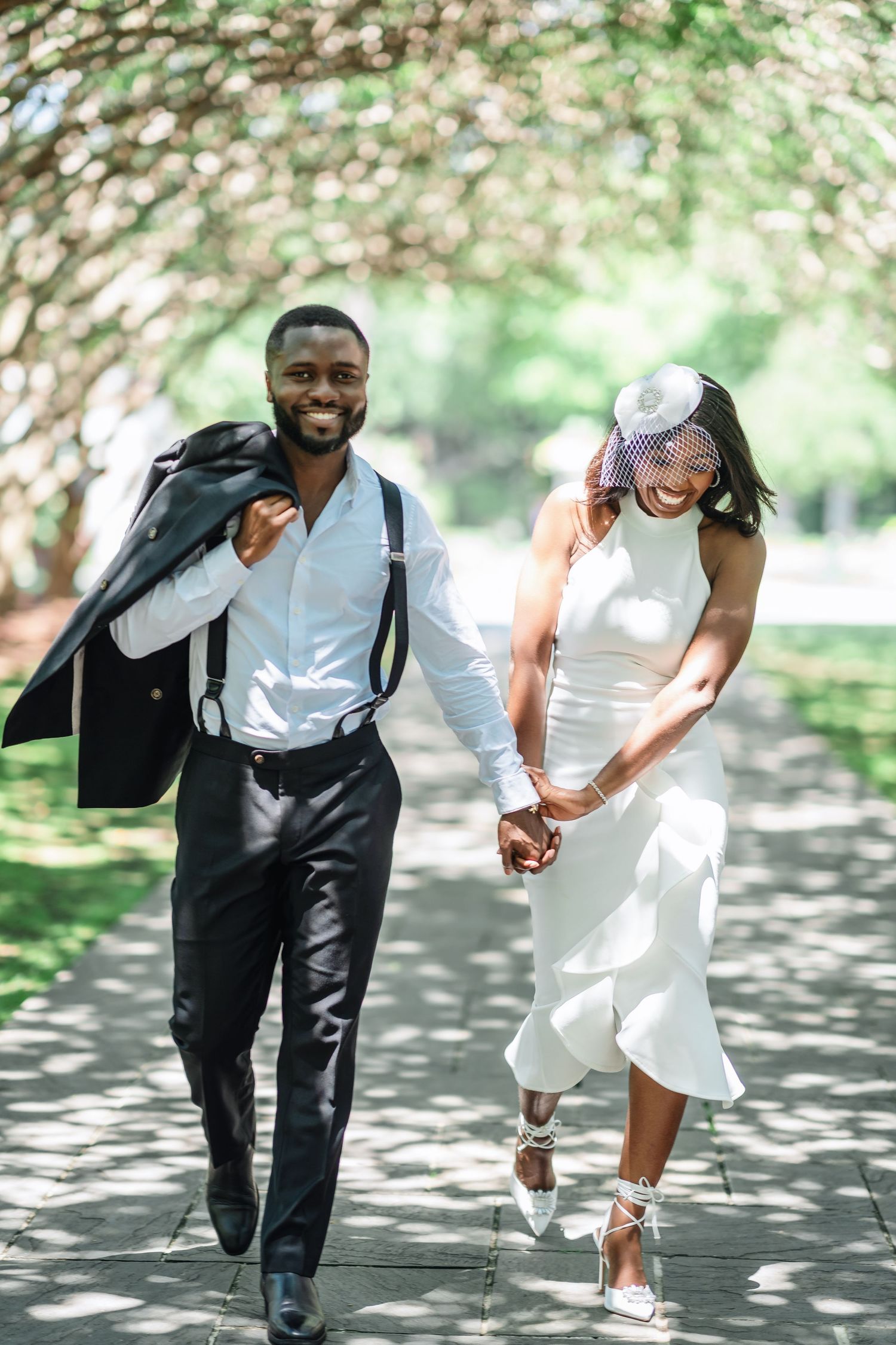 Playful couple in formal attire runs and poses along a tree-lined path with dappled sunlight.
