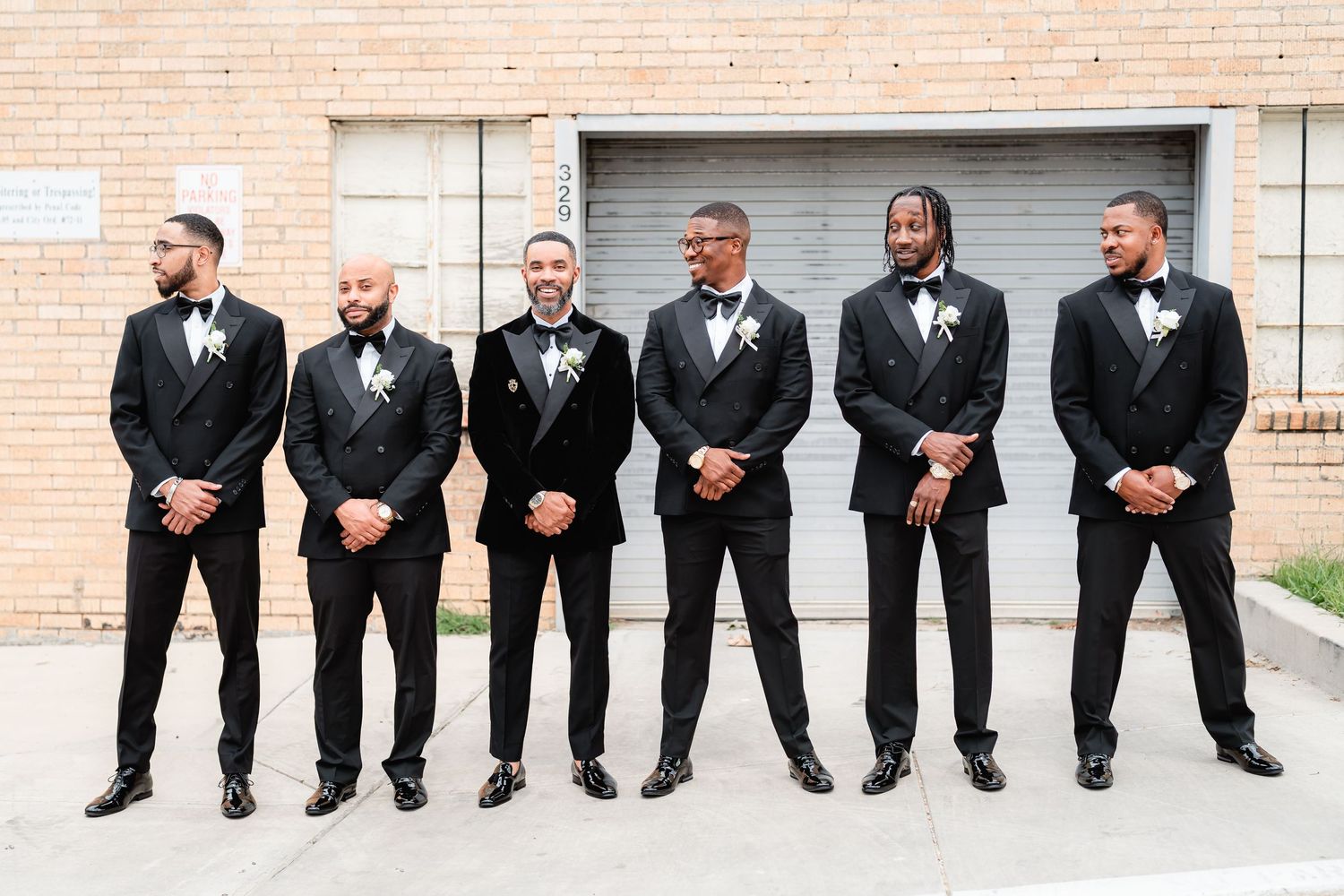 Row of groomsmen in black tuxedos sitting on brick wall showing their dress shoes and socks.