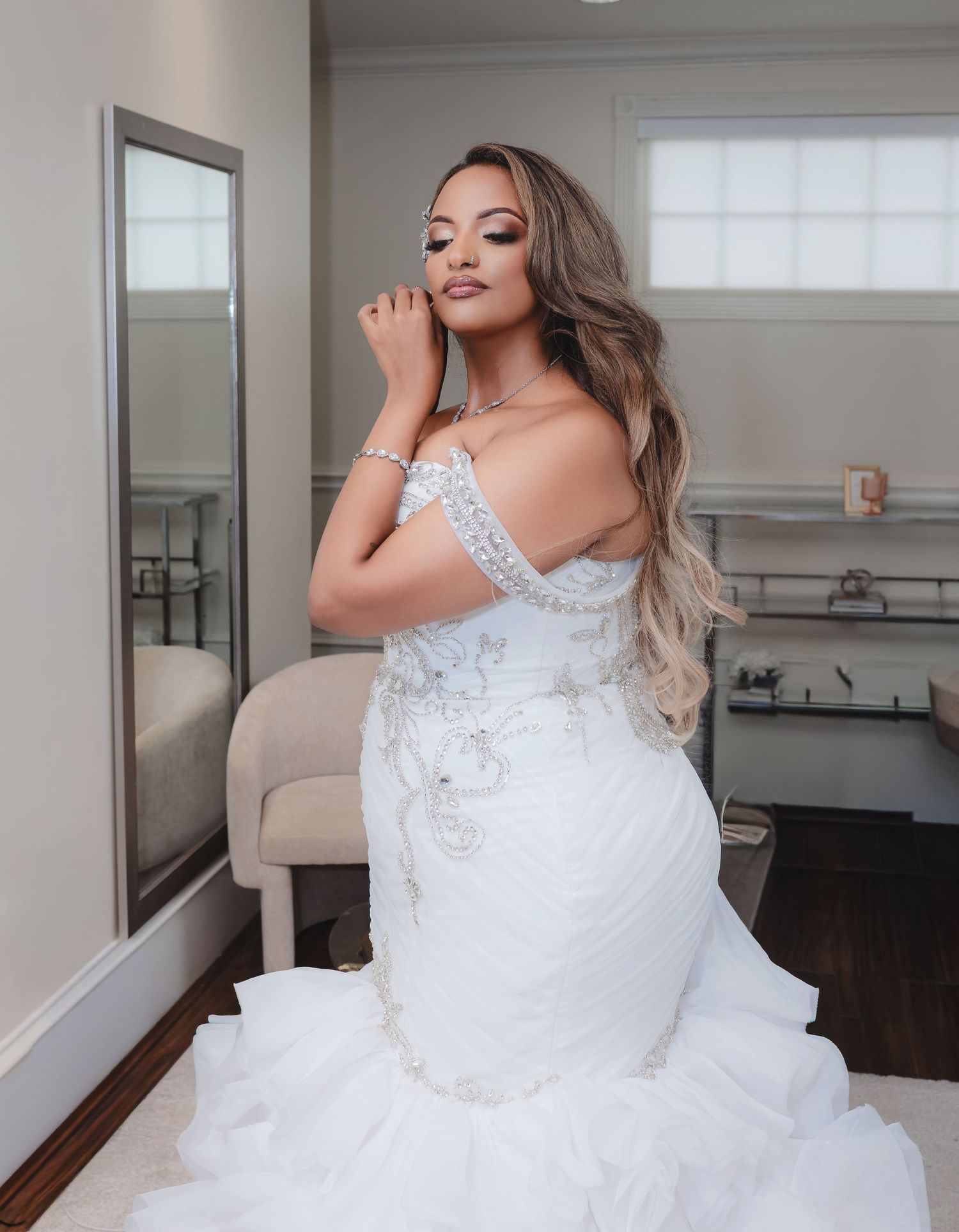 A bride poses in an embellished white wedding gown with long wavy hair in an elegant indoor setting.