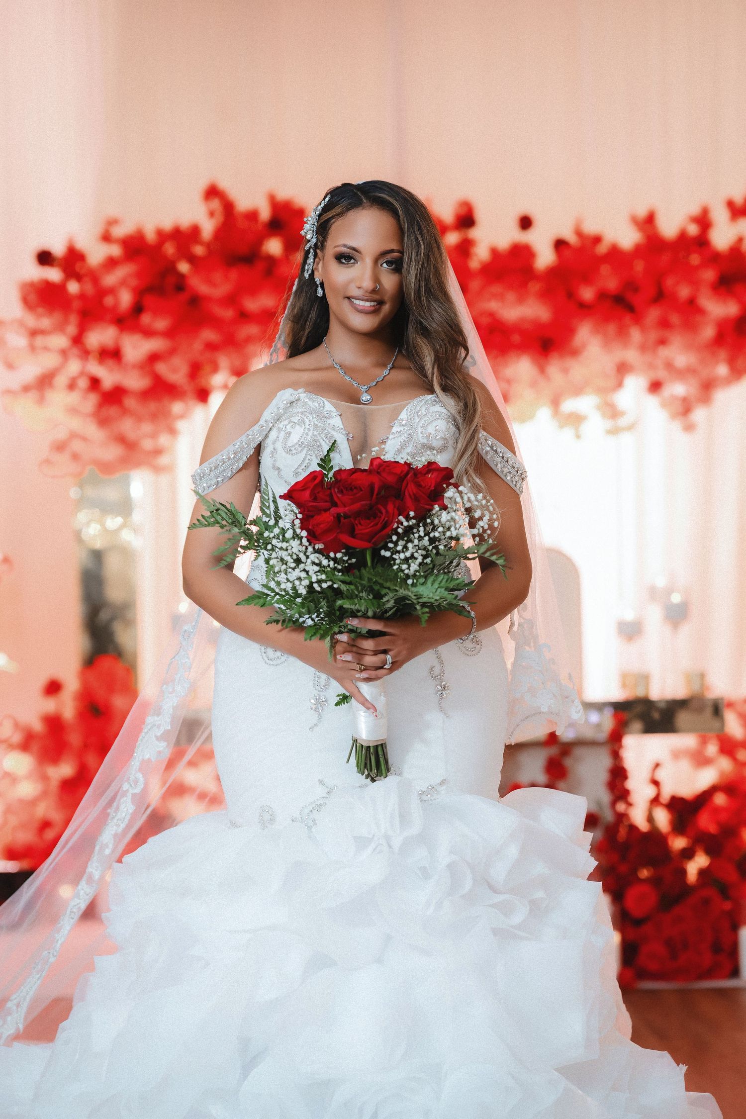 A bride in a white wedding dress holds a bouquet of red roses against a backdrop of red floral decorations.