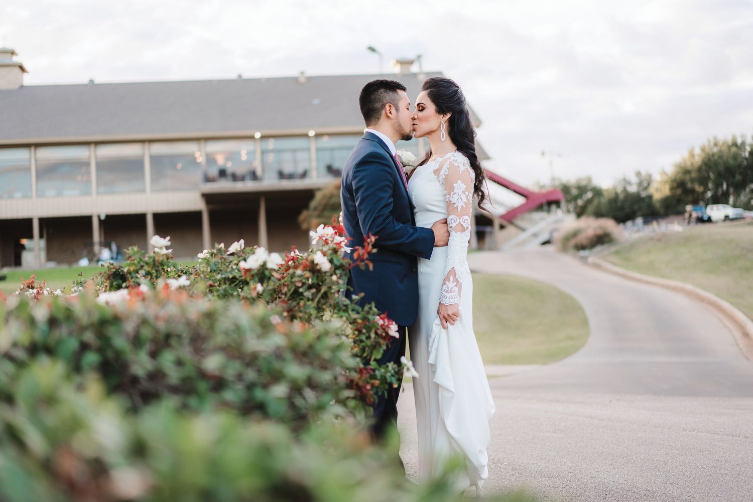 A couple shares a romantic moment on a winding pathway with greenery and buildings in the background.