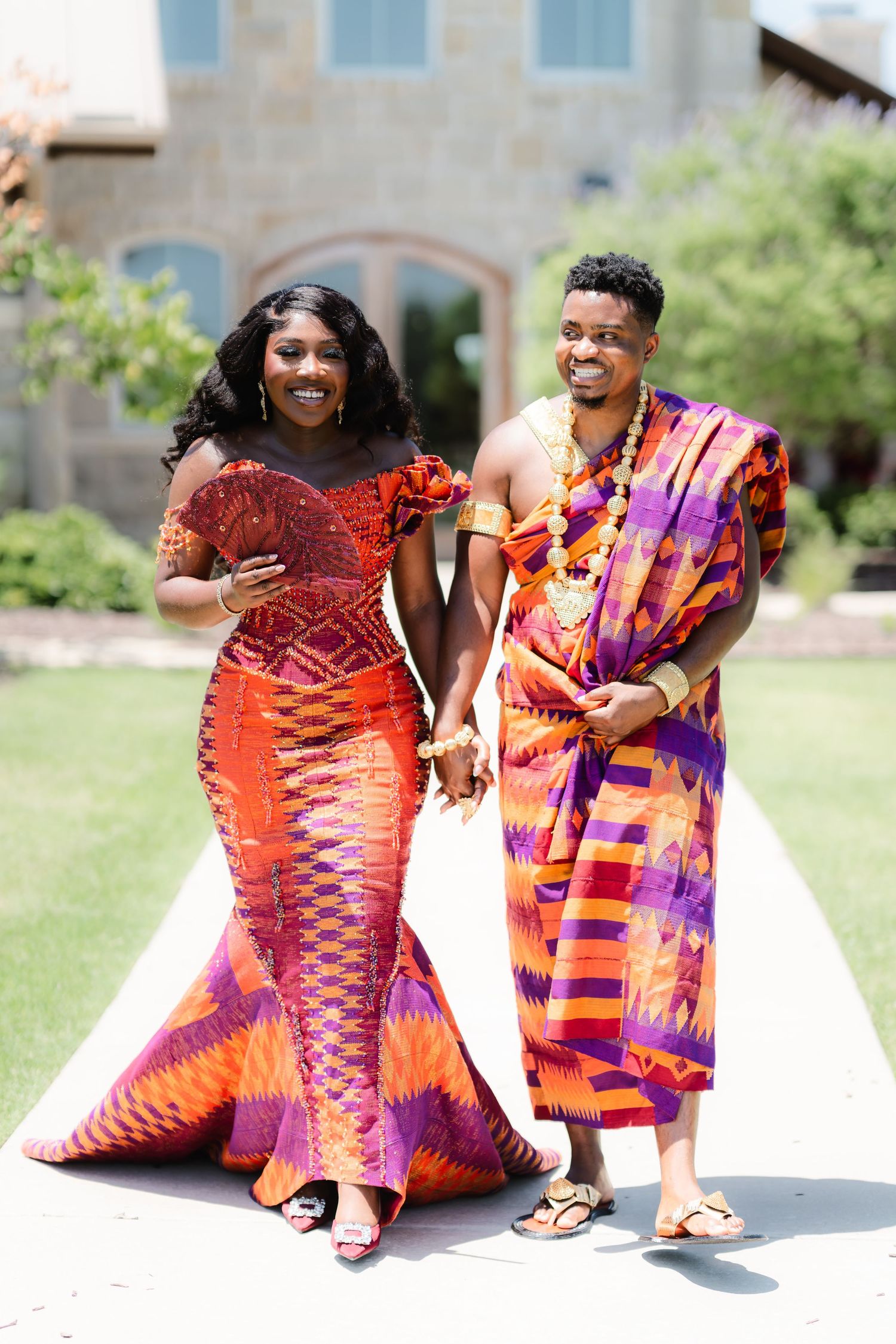 A couple wearing vibrant traditional African attire poses together on a grassy lawn in front of a building.