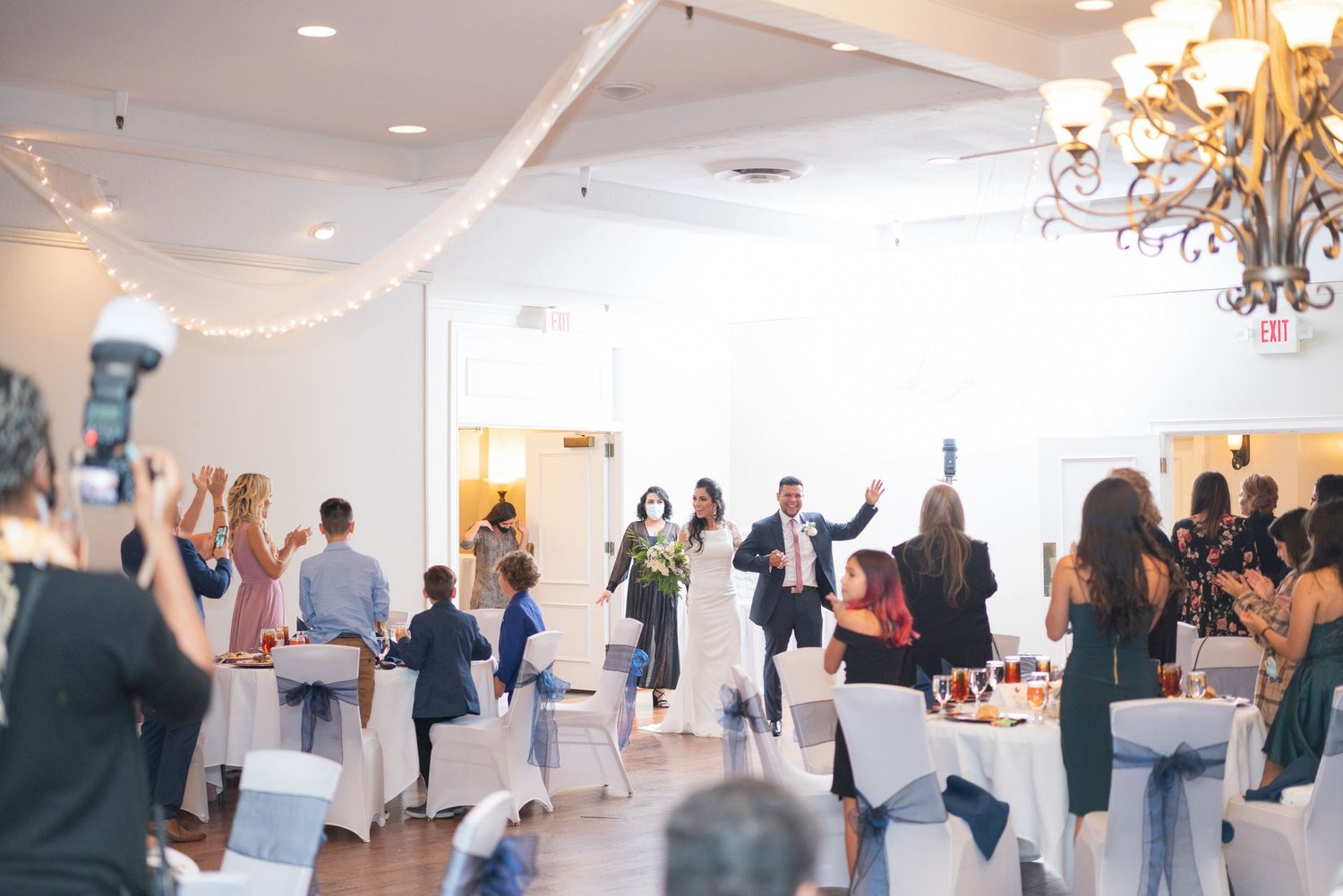 Wedding reception venue decorated with white draping and chandeliers as guests mingle.