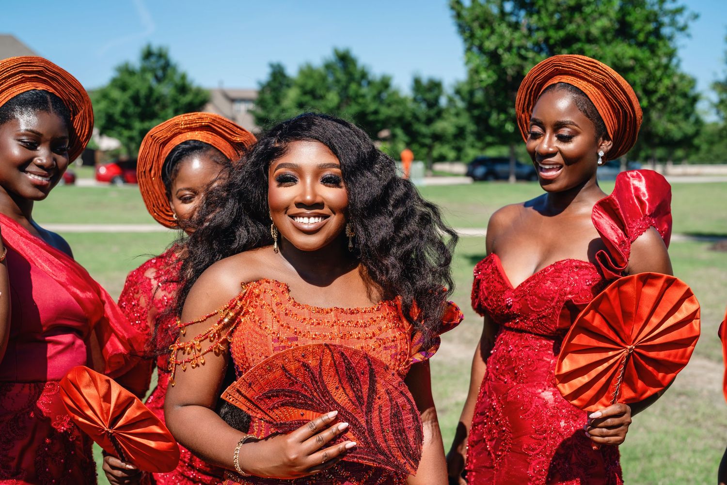 Wedding party members in vibrant red dresses and orange accessories pose together outdoors on a sunny day.