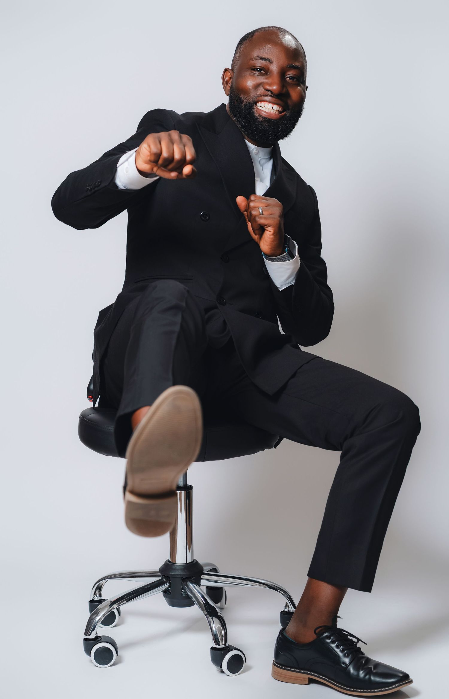 Professional in black suit playfully poses on office chair against white background showing dynamic movement.