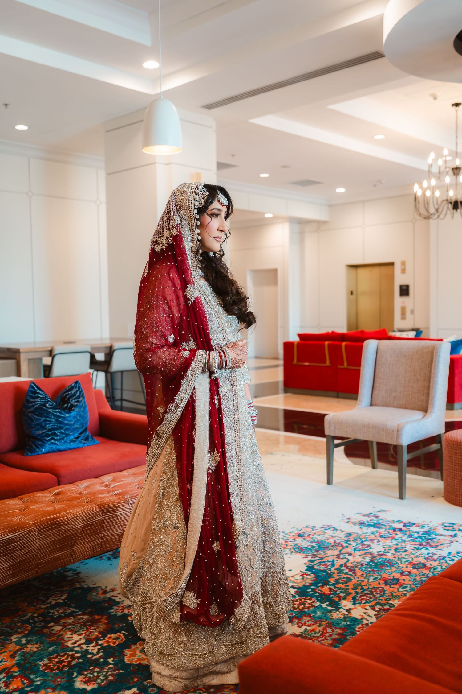 A bride in an ornate red and gold lehenga poses in a modern interior with red seating.