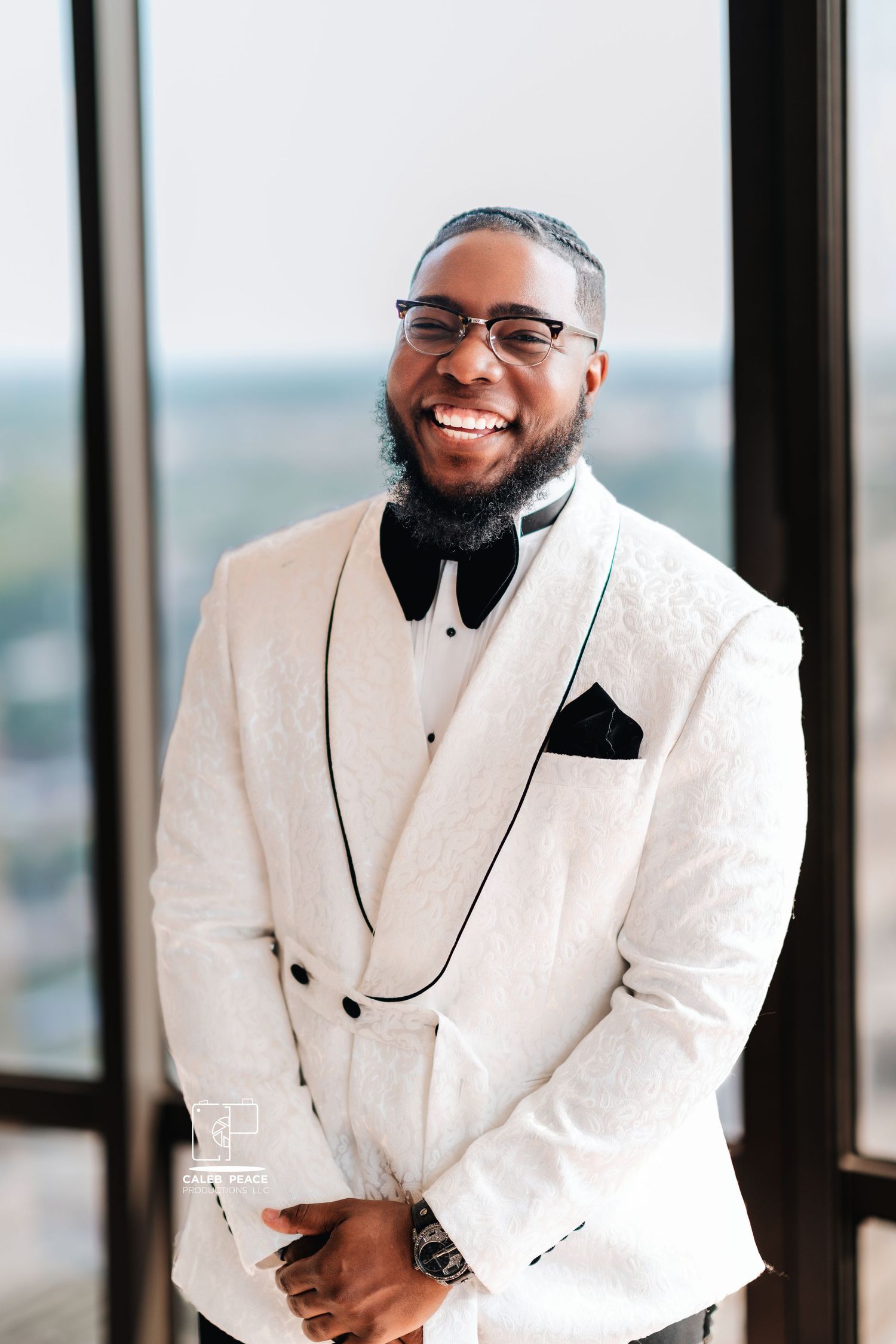 A groom wearing a white tuxedo jacket and bow tie smiles warmly by a window.