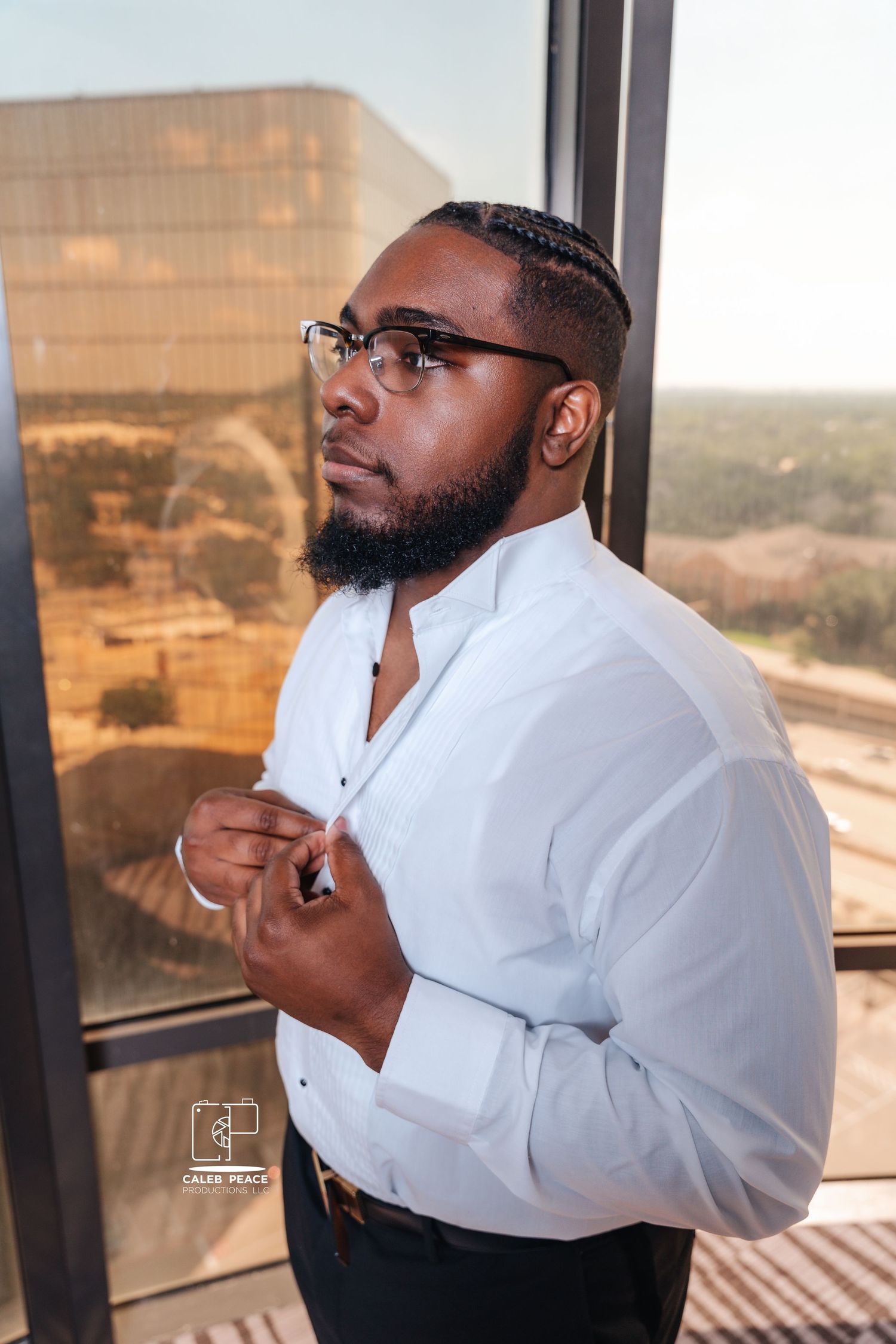 Groom adjusting his white dress shirt while looking out window in preparation for wedding.