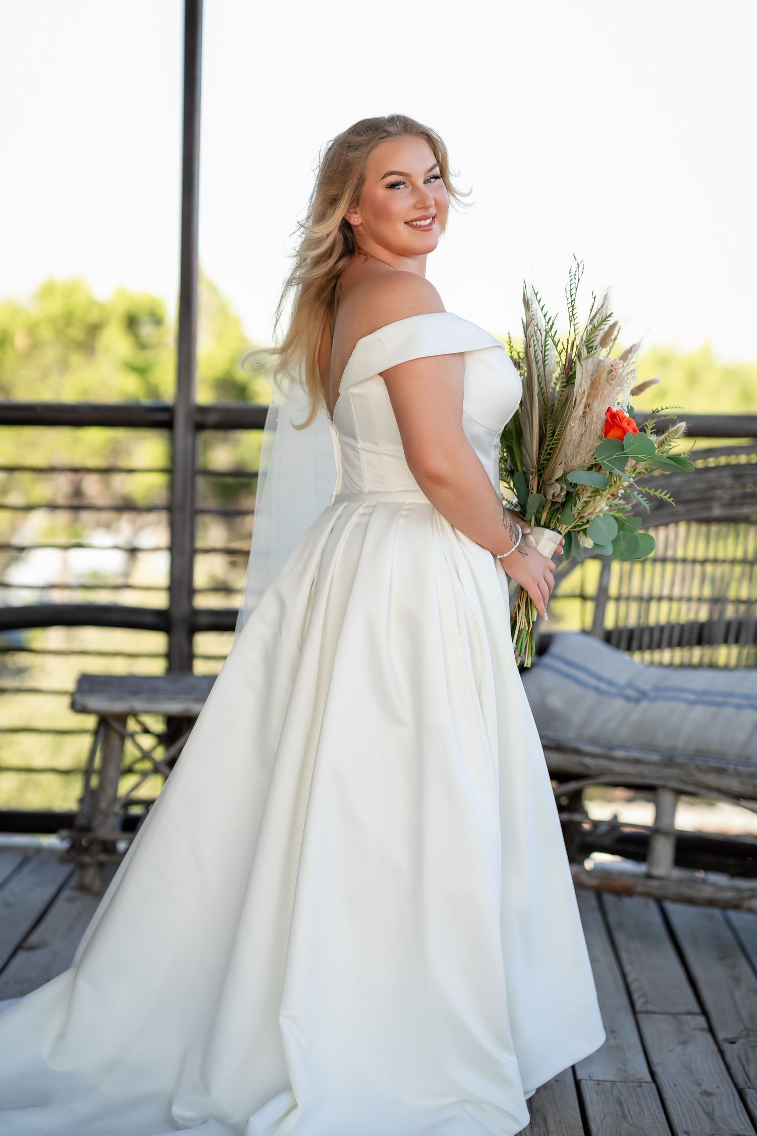 Bride in flowing white wedding dress poses on wooden deck with scenic backdrop.