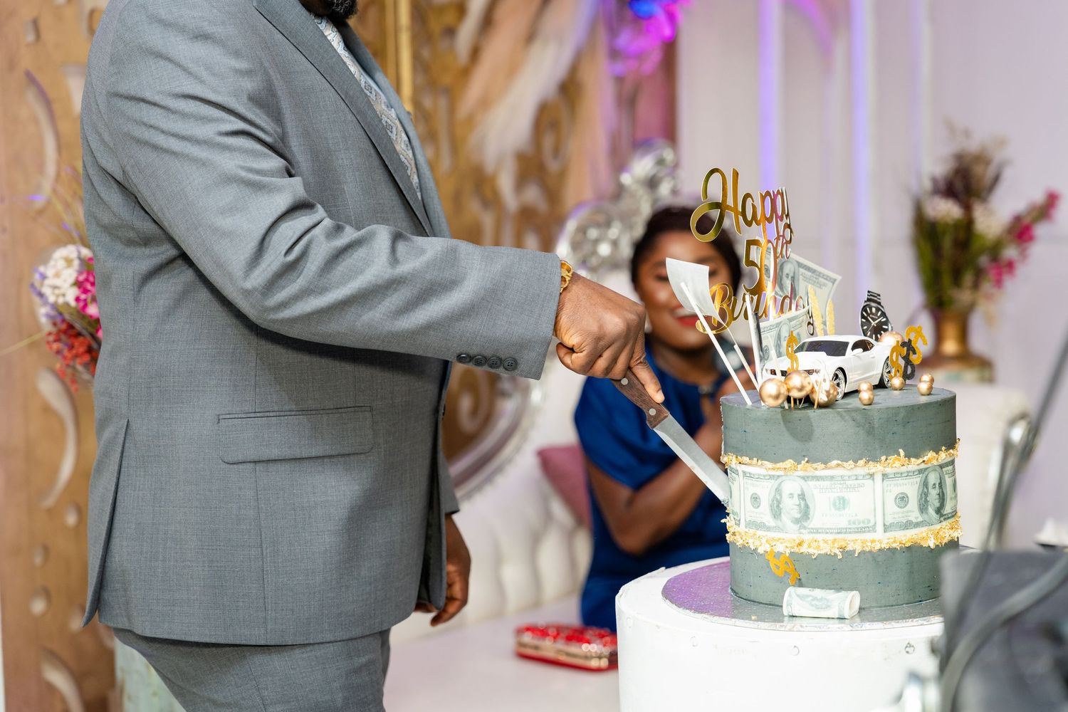 A wedding reception scene with guests gathering around a tiered white cake decorated with flowers and lights.