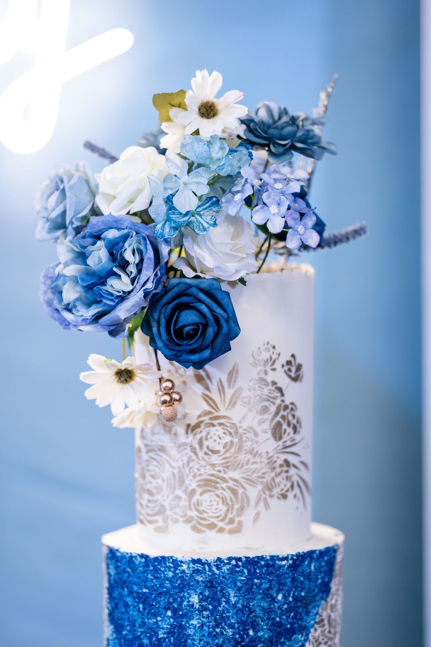 Elegant white and royal blue wedding cake decorated with floral details and sparkling sequins against blue background.