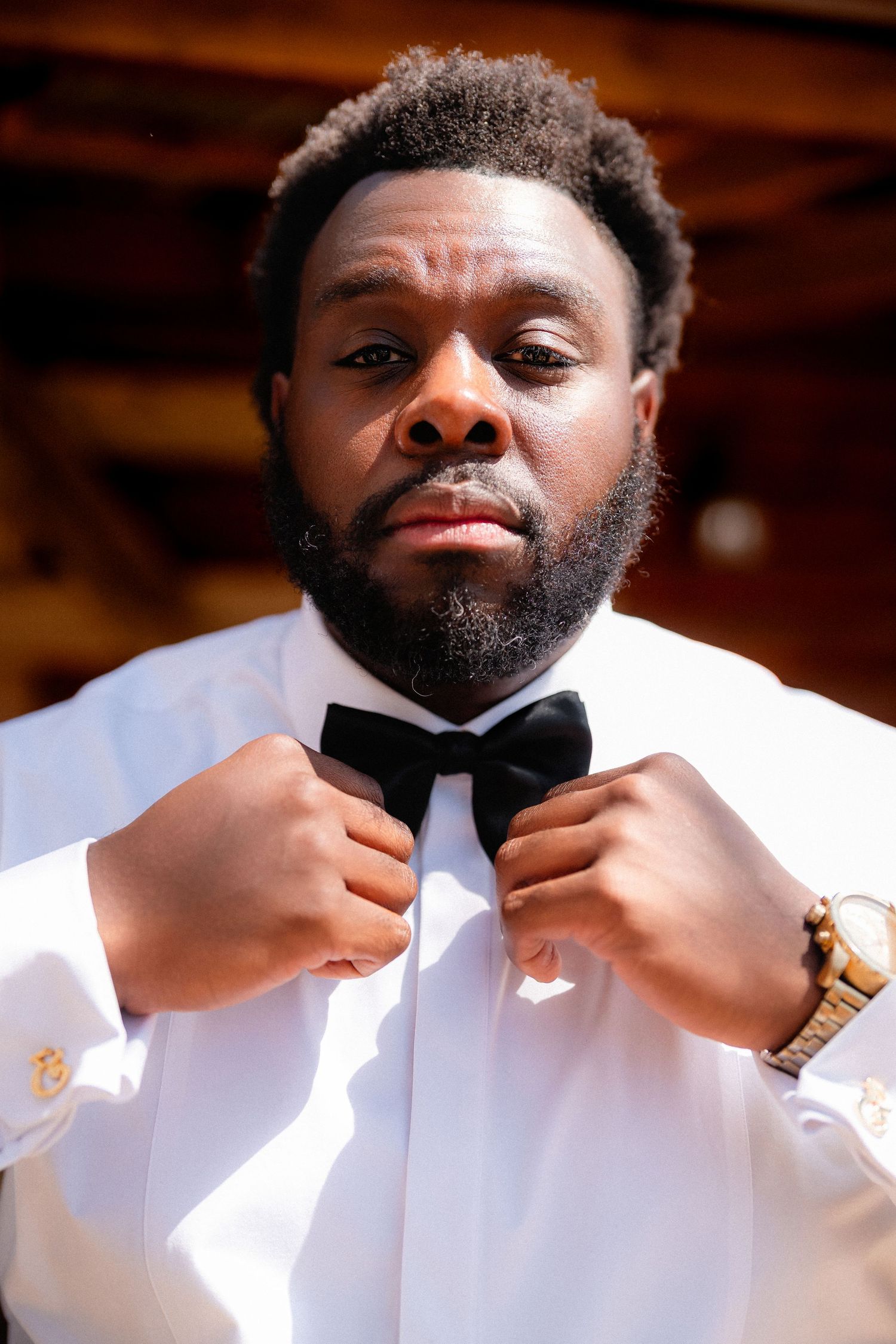 A groom adjusts his black bow tie while wearing a crisp white tuxedo jacket and gold watch.